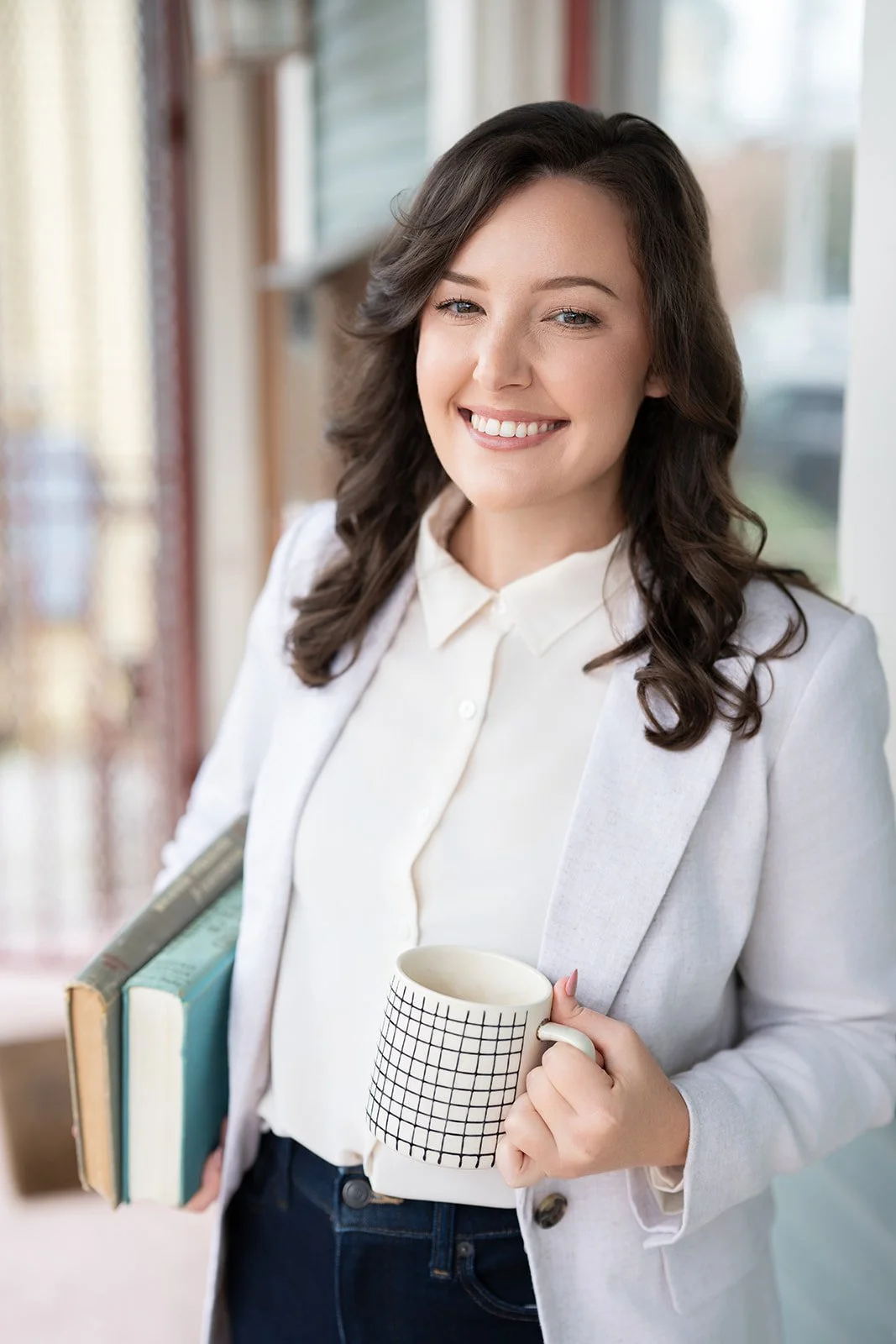 Victoria Rodriguez, Licensed Professional Counselor and PhD in Counselor Education, smiling in a light gray blazer while holding books and a mug by a window