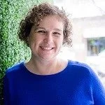 Sarah Erdman, Associate Professional Counselor, smiliing in a bright blue crewneck top standing next to a green plant wall