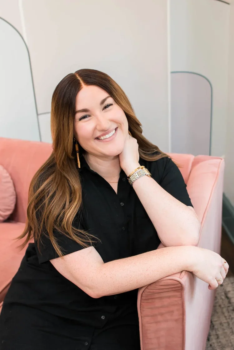 A woman with long brown hair smiling and sitting on a pink sofa, resting her head on her hand, wearing a black shirt and gold jewelry.