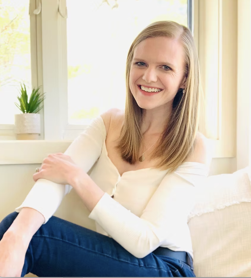 Dr. Jessica Rabon, a licensed clinical psychologist, smiling in a white off-the-shoulder long-sleeved top, dark jeans, and a pendant necklace seated indoors next to a bright window with a small potted plant