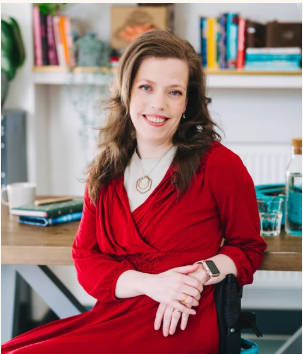 Grace Quantock, psychotherapeutic counsellor and writer, smiling wearing a red dress, sitting in front of a desk with bookshelves behind her