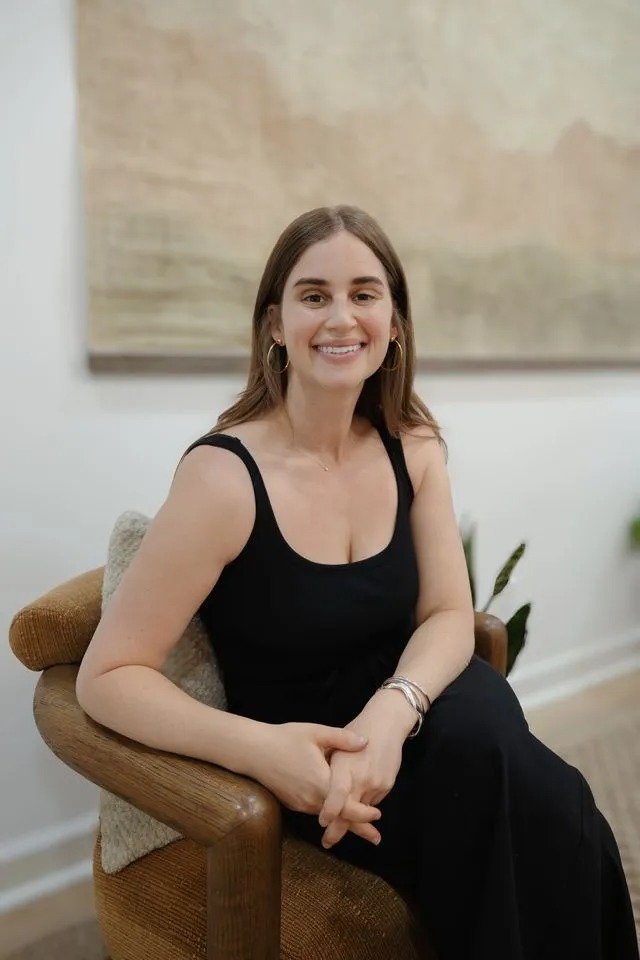 Meg Bearman, therapist and art therapist, smiling in a black sleeveless dress and gold hoop earrings seated in a wooden armchair