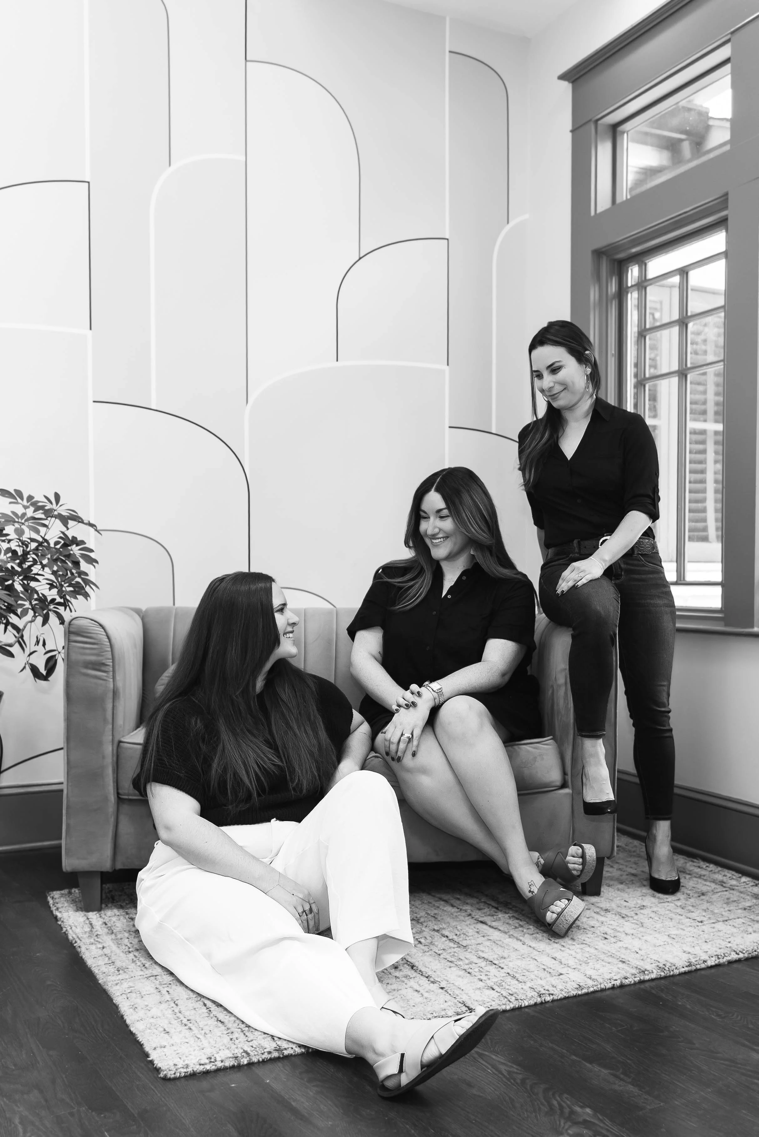 A group of five women in a living room, with some seated on a sofa and one standing by the window, engaging in conversation and smiling. The room has large windows, decorative wall panels, and a rug on a wooden floor.