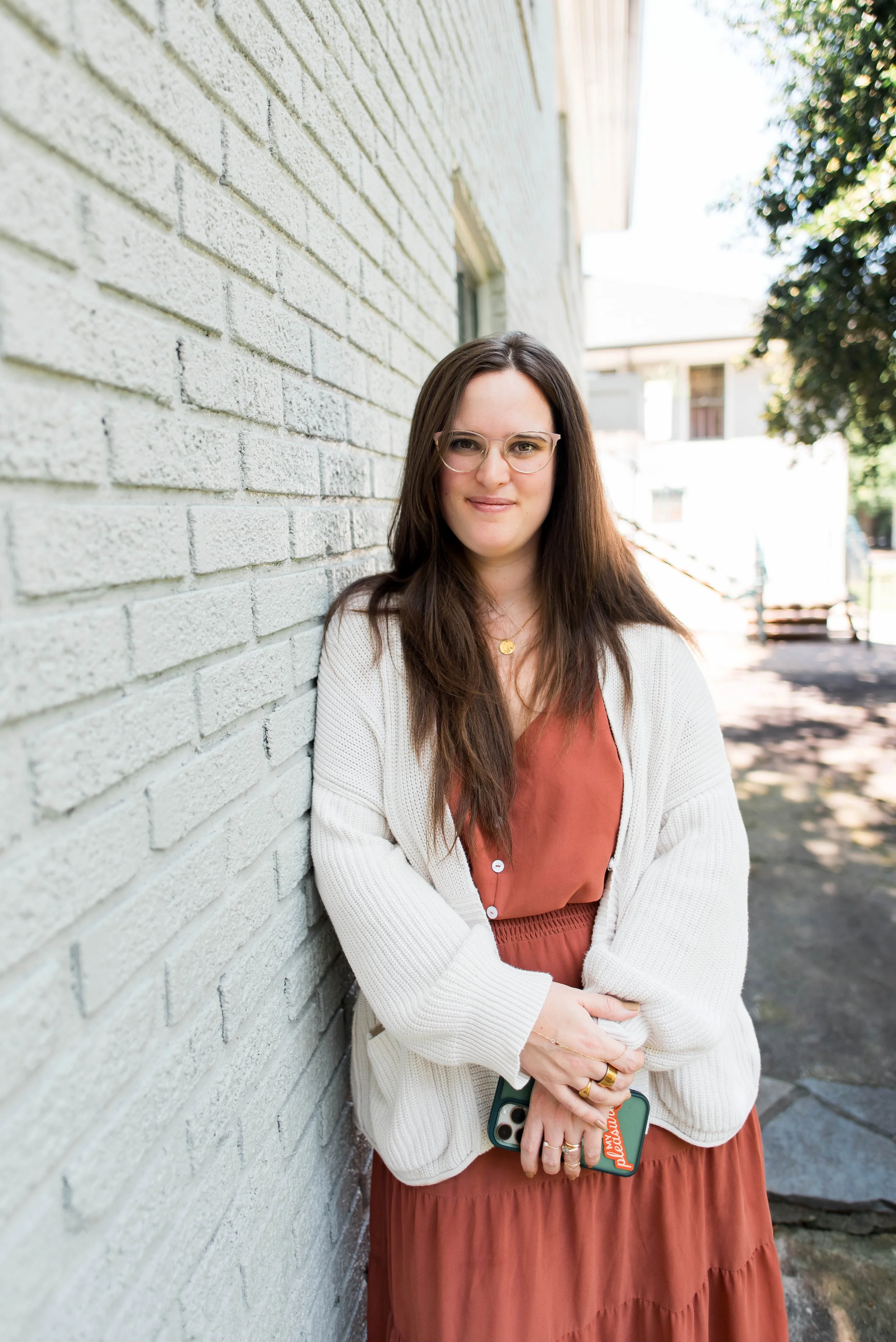 A young woman with long brown hair, glasses, and a gold necklace smiling while leaning against a white brick wall outdoors. She is wearing a rust-colored dress and a cream cardigan, holding a smartphone with a colorful case. Sunlight and trees are visible in the background.