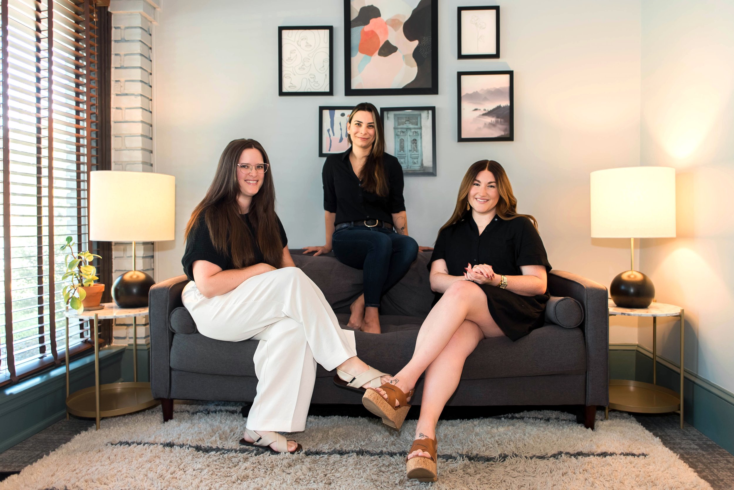 Three women sitting on a gray couch in a cozy living room with gallery wall art behind them. One woman is seated on the left, wearing glasses and a black top with white pants. The second woman is seated on the right, wearing a black dress. The third woman is sitting on the back of the couch, wearing a black top and jeans. There are two lamps, one on each side of the couch, and a window with wooden blinds on the left.