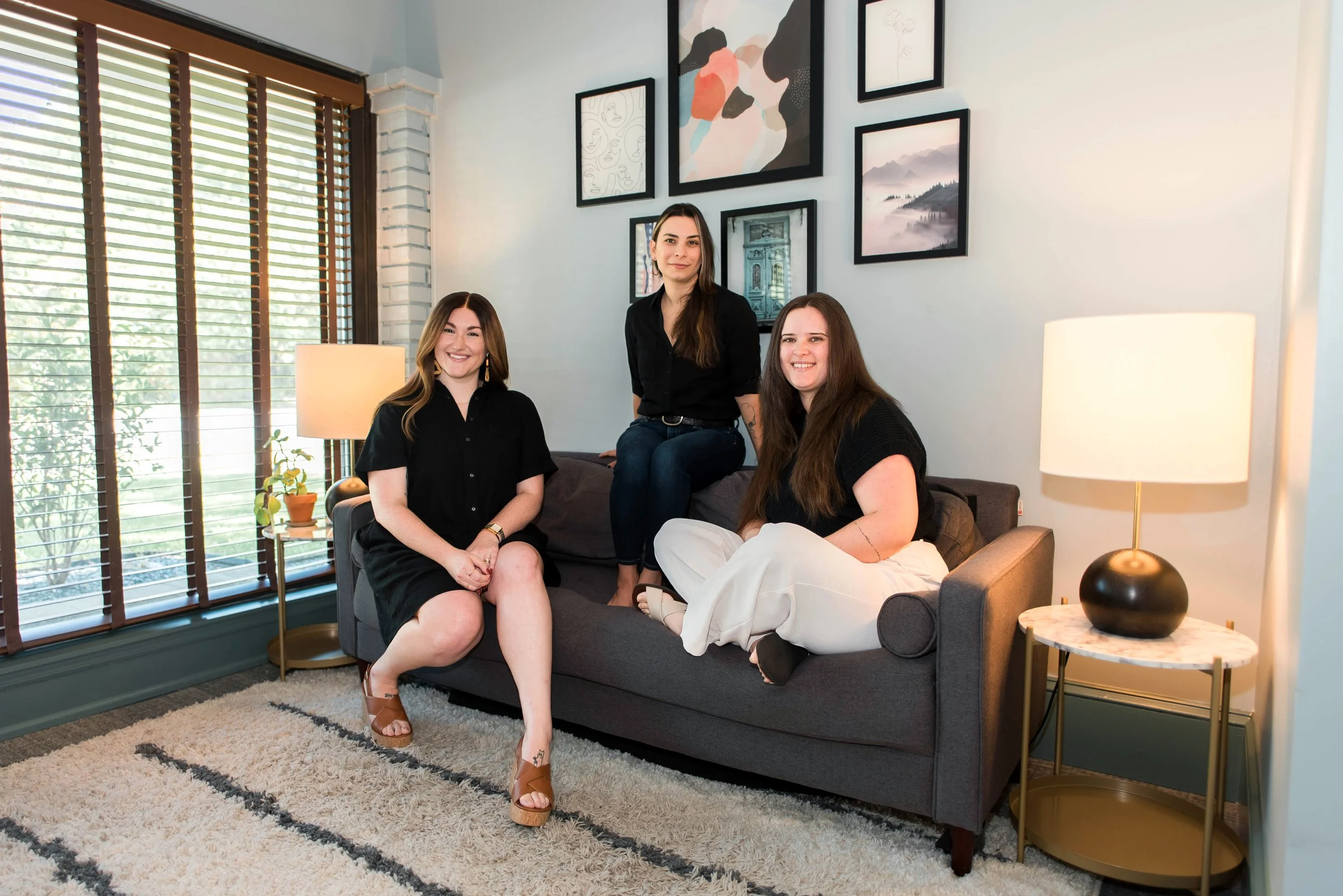 Four women sitting and standing on a gray sofa in a cozy living room with framed artwork, blinds, a table lamp, and a plush beige and black rug.