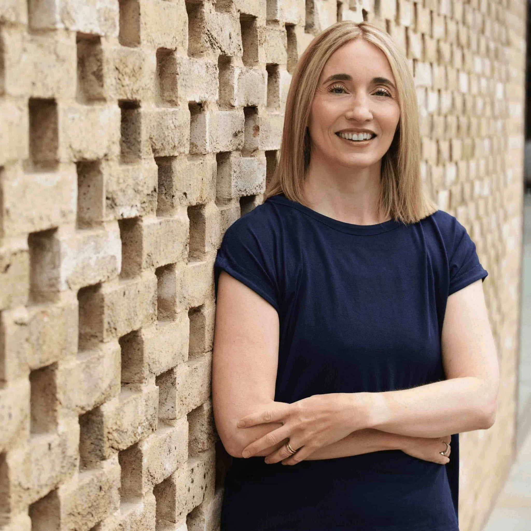 Jeannie Di Bon, Movement Therapist, smiling wearing a navy blue t-shirt, leaning against a patterned brick wall with her arms crossed