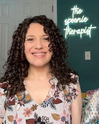 Nadia Clontz, Licensed Professional Counselor, smiling in a floral patterned top in front of a neon sign that reads "the spoonie therapist"