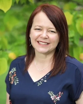 Stephanie Rotenberg Lewis with auburn hair wearing a navy V-neck shirt in front of green leafy foliage.