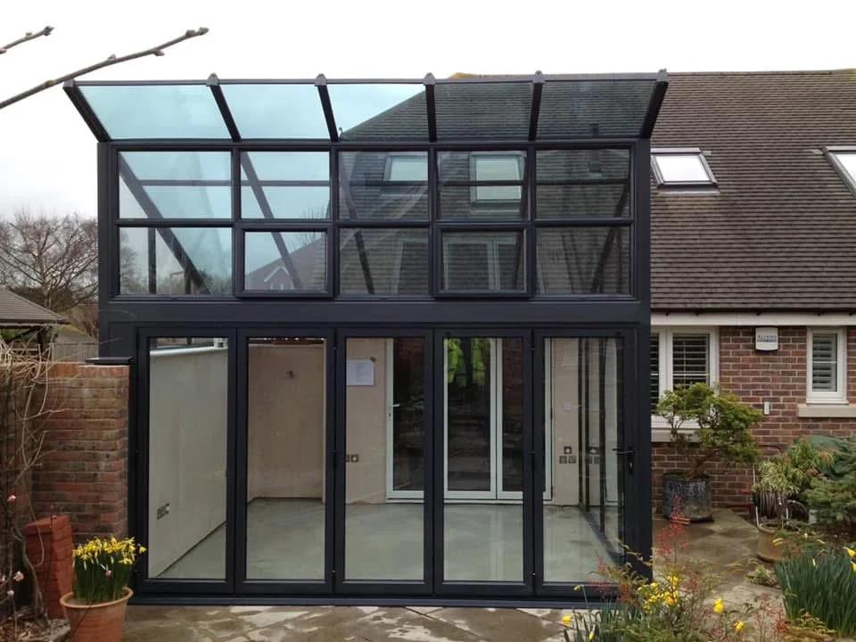 A modern glass extension with a black frame attached to a brick house, featuring large glass windows and doors, with a small garden area in front.