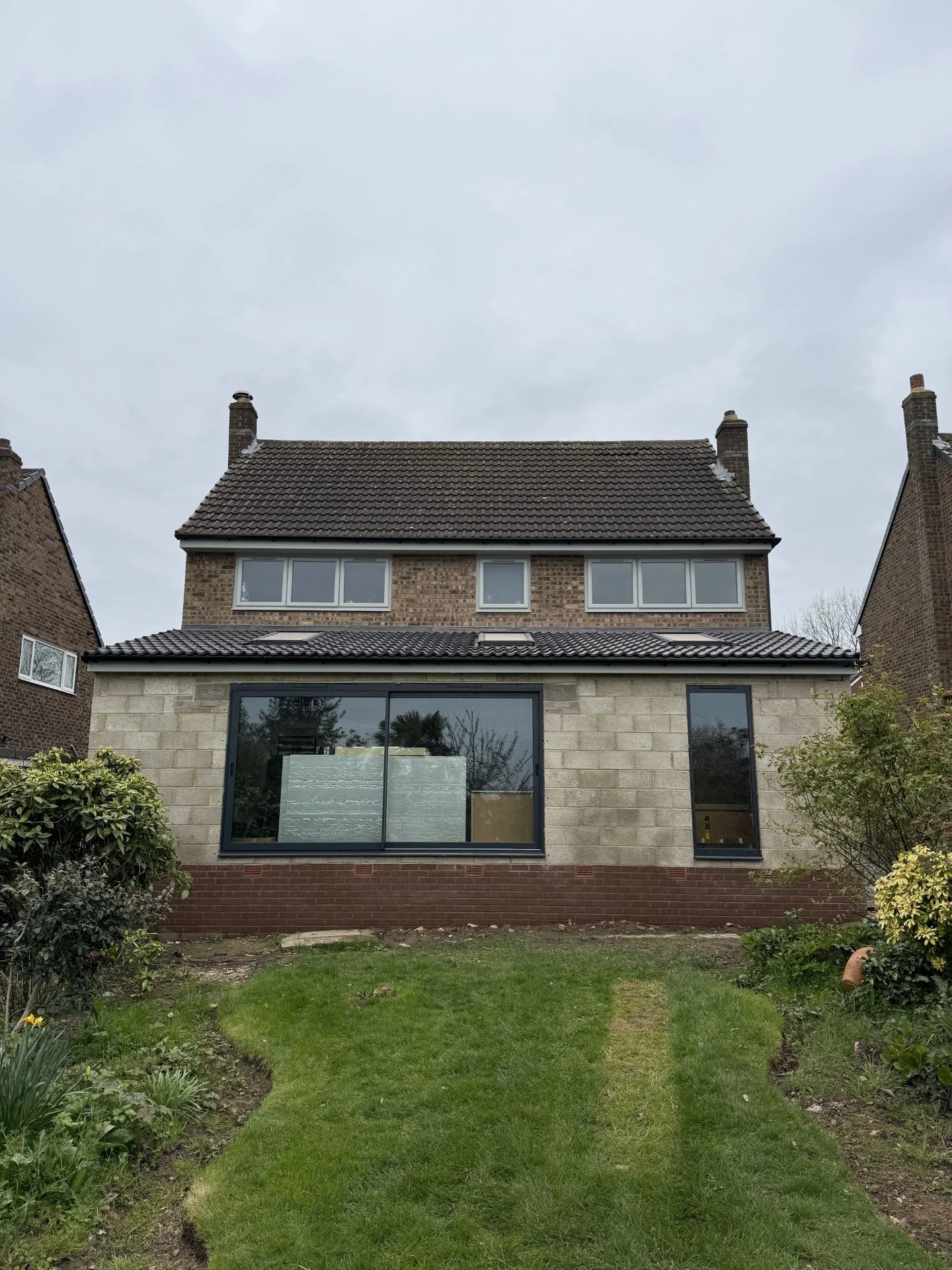 Front view of a multi-story house with brick and stone exterior, large glass windows, and a small front yard with grass and shrubs, under a cloudy sky.