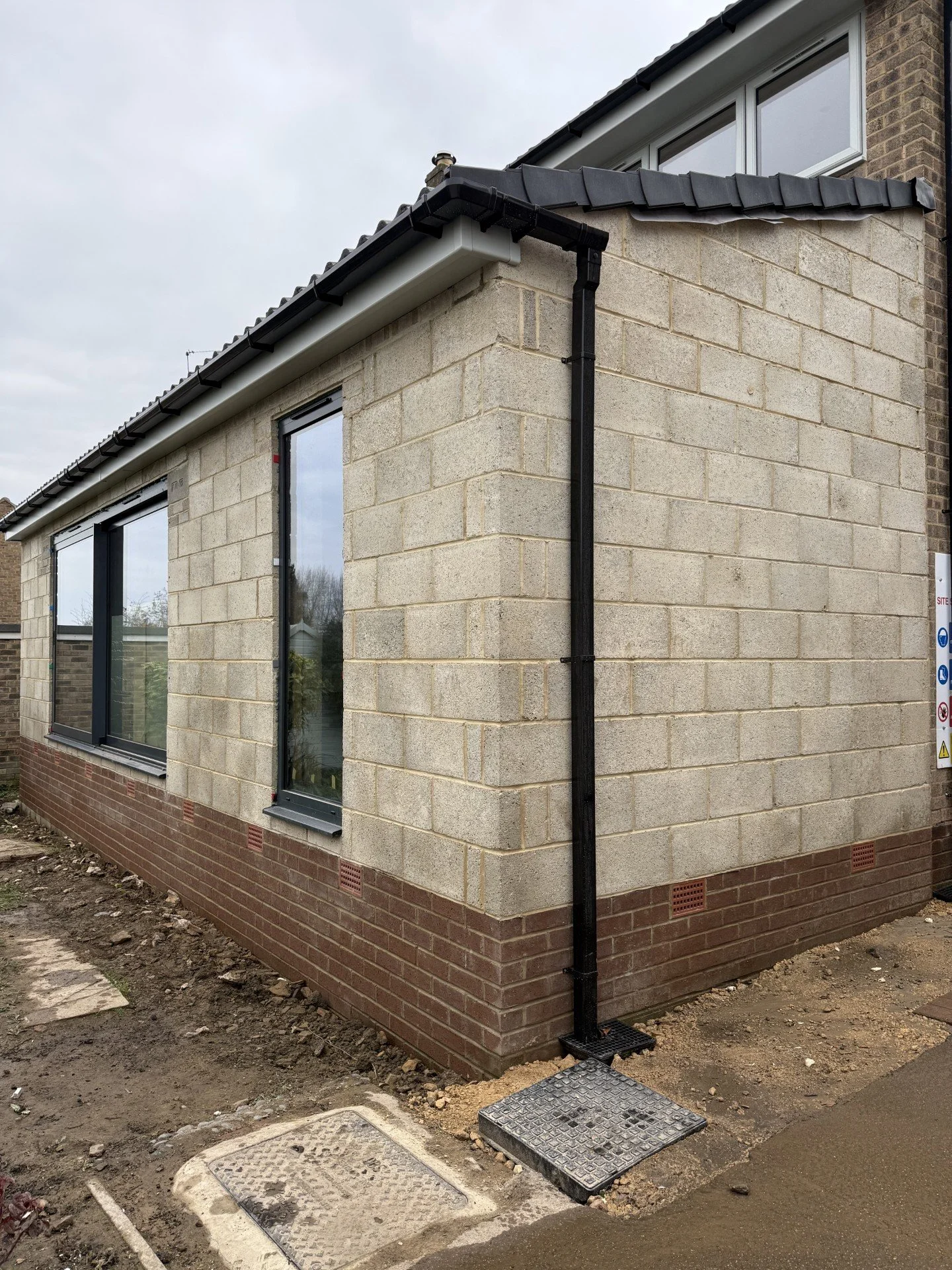 A newly constructed house with beige brick walls, black window frames, and a black gutter system, with a muddy ground and a small paved area near the corner.