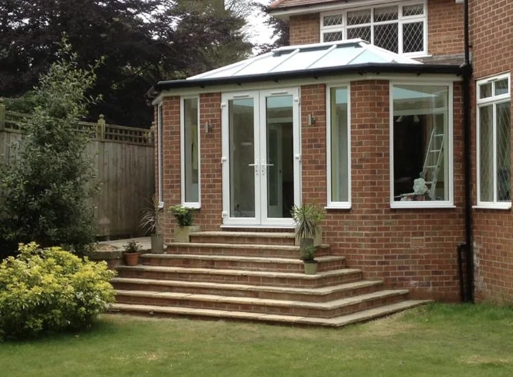 Backyard view of a brick house with a conservatory extension and wooden steps leading to glass doors.