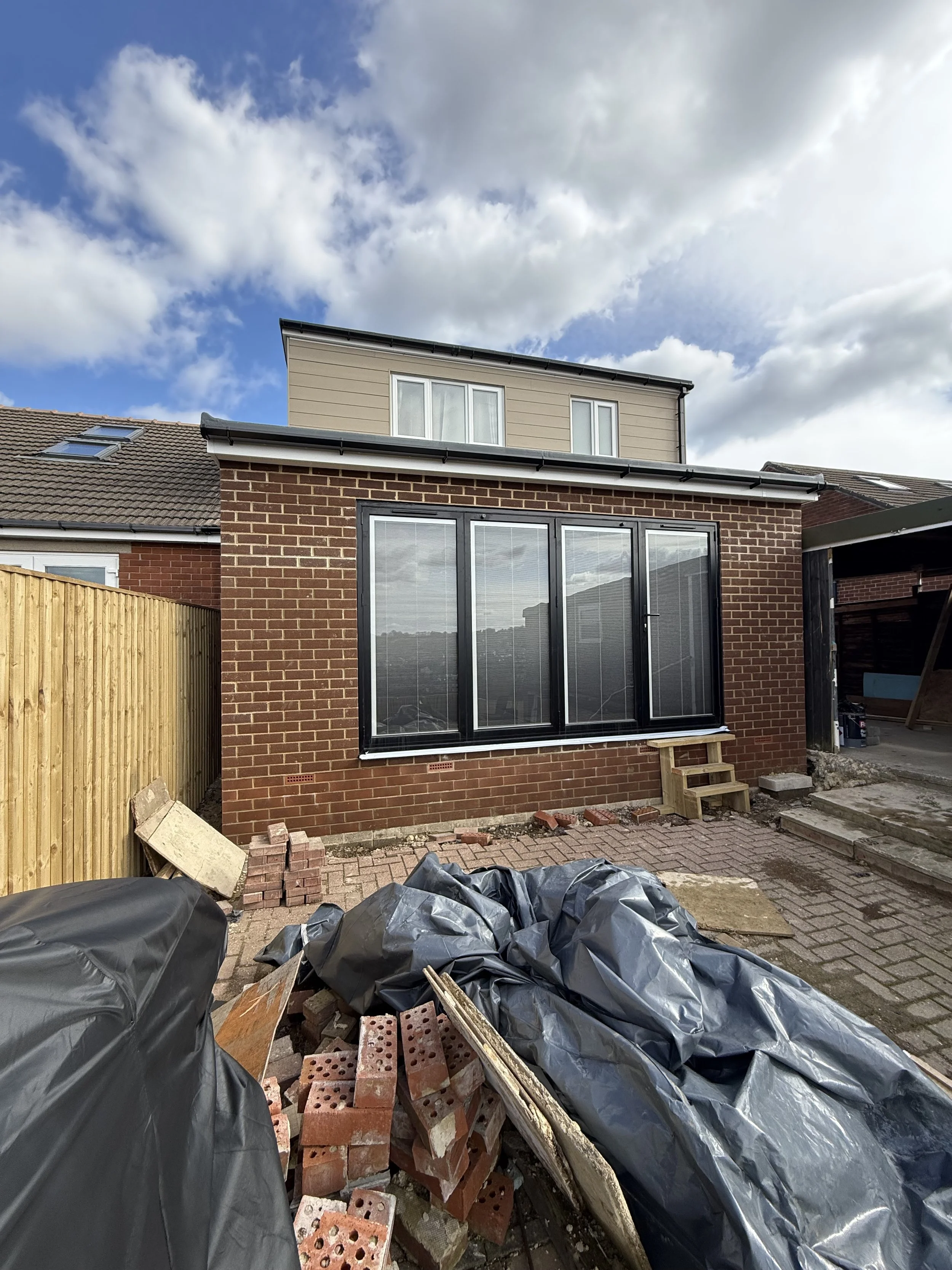 Backyard exterior of a brick house under renovation with construction materials, bricks, covered with black plastic, a small wooden staircase, and a paved patio area under a partly cloudy sky.
