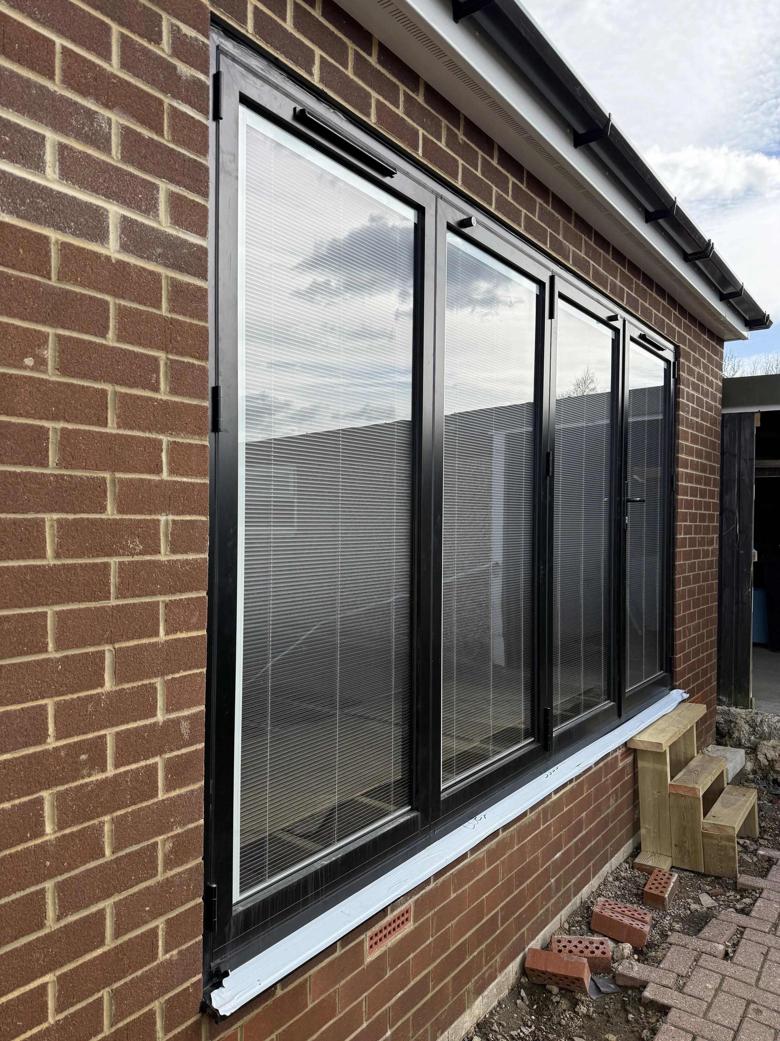 A large window with black metal frames and glass panes installed on a red brick exterior wall of a building. The glass reflects the sky and clouds. There is a small wooden step at the base of the window and some bricks and pavers on the ground nearby