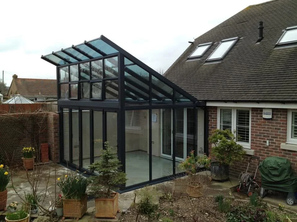 A glass extension attached to a brick house with sliding glass doors and a sloped roof with skylights, surrounded by a garden with potted plants and gardening tools.