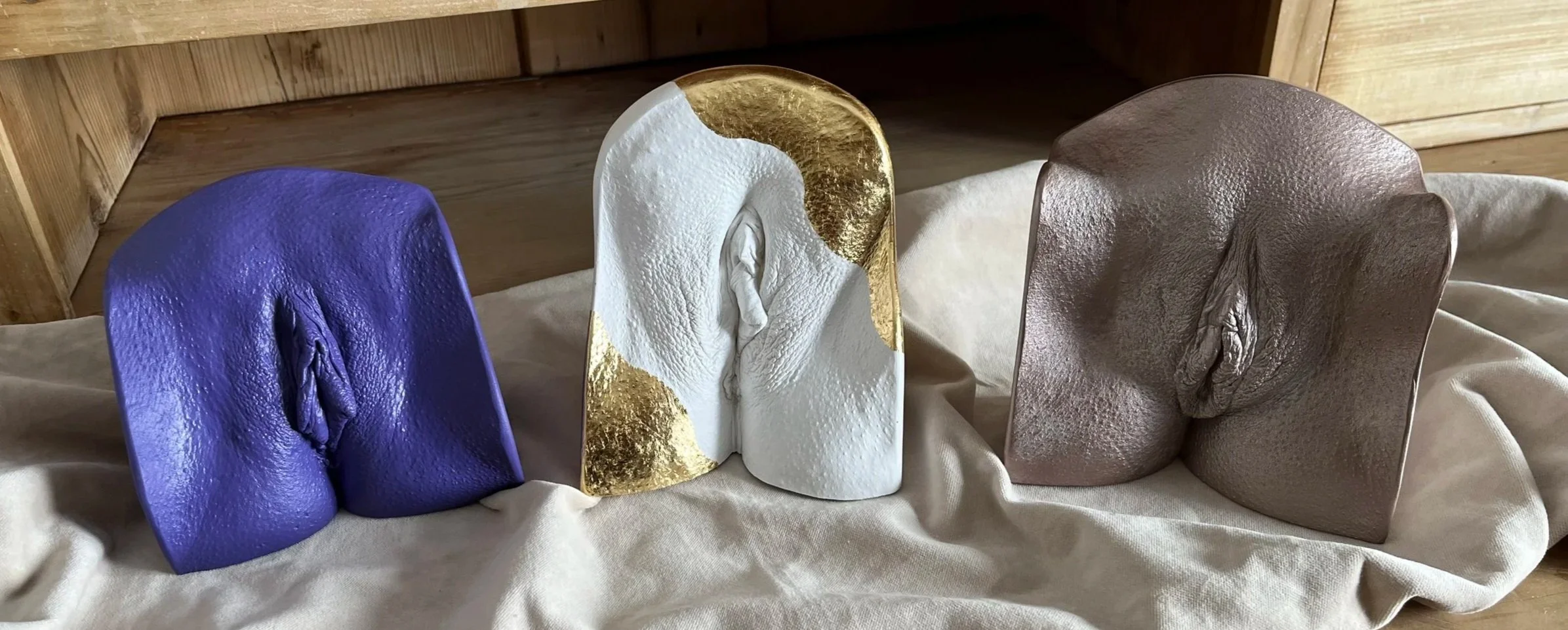 Drei Vulvaabformungen in den Farben Lila, Weiß mit Goldakzenten und Braun.
