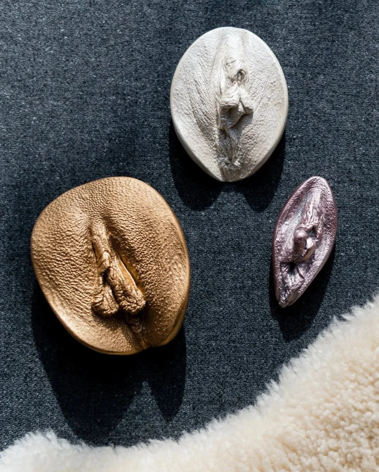 Drei Mini Muses (detailgetreue Abformungen von weiblichen Genitalien). Die Skulpturen sind in unterschiedlichen Farben: gold, perl-weiß und metallic rosé, auf einer schwarzen Oberfläche ausgestellt.