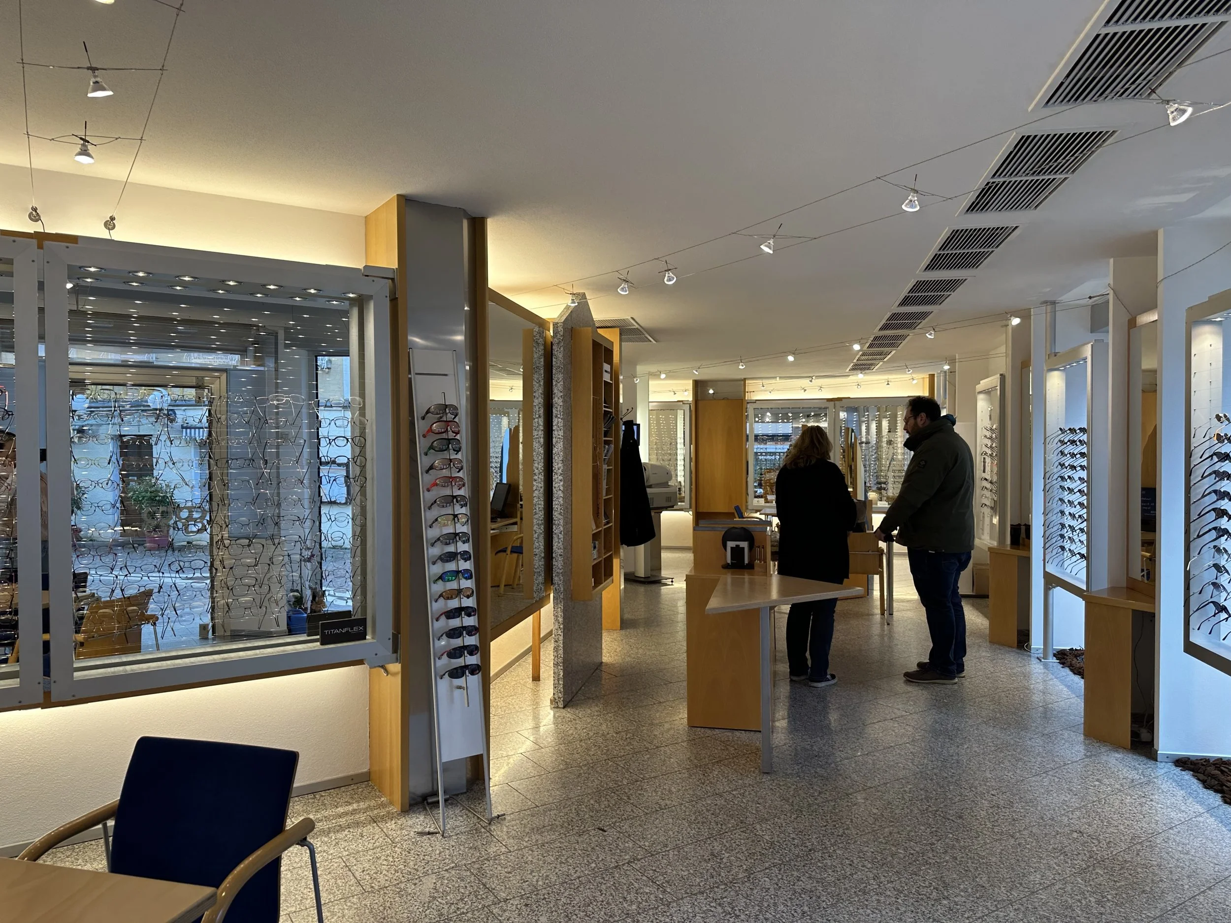 Optikergeschäft (Bammert Brillen in Aalen) mit Kunden, die Brillen auswählen, Regale mit Brillen und Sonnenbrillen, große Fenster mit Blick auf die Straße, helle Innenbeleuchtung.