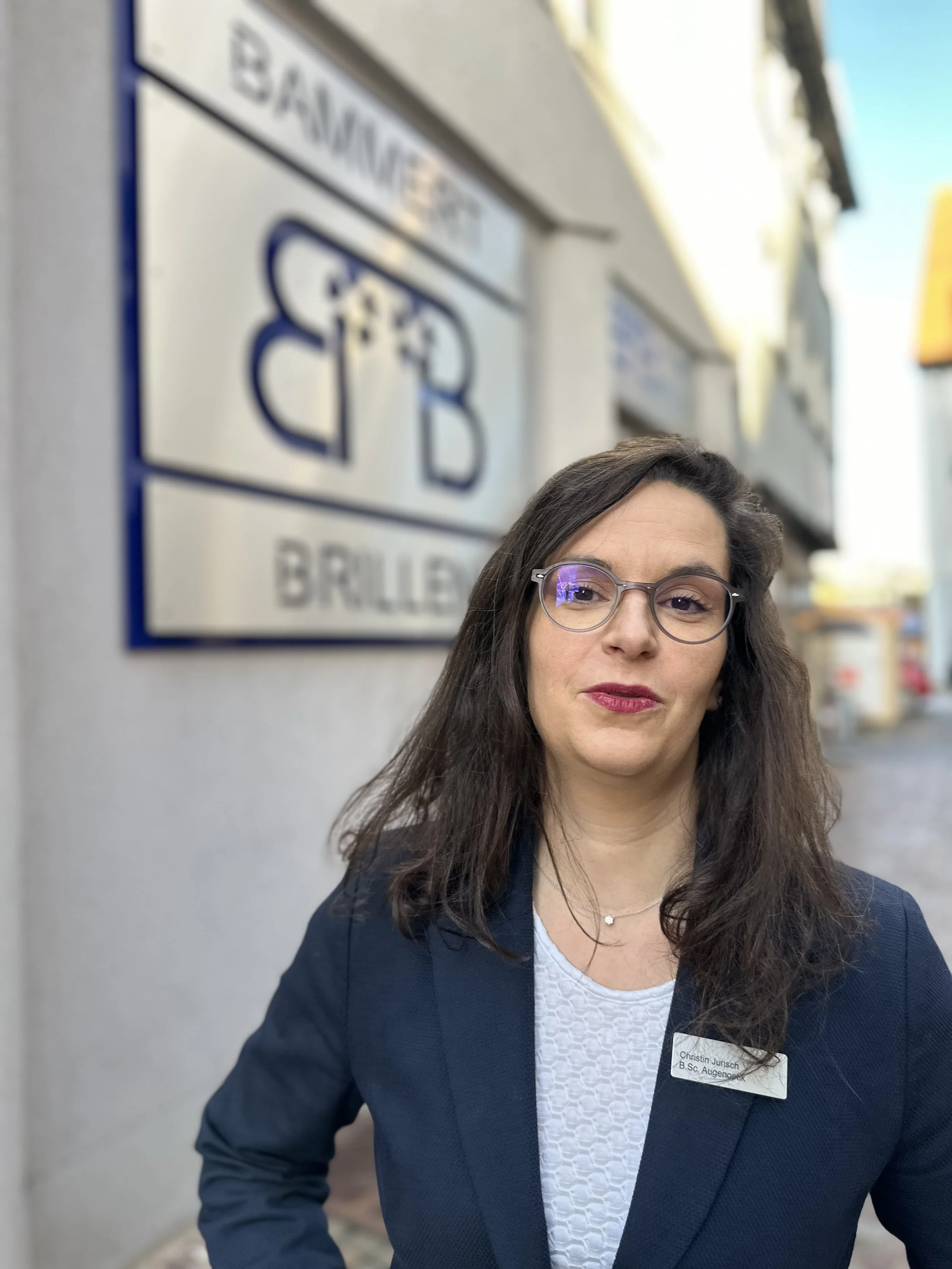 Frau (Chef)  mit Brille, dunklem Haar und Blazer vor einem Geschäft mit einem blauen und weißen Schild, das 'BB' und 'Bammert Brillen' zeigt.