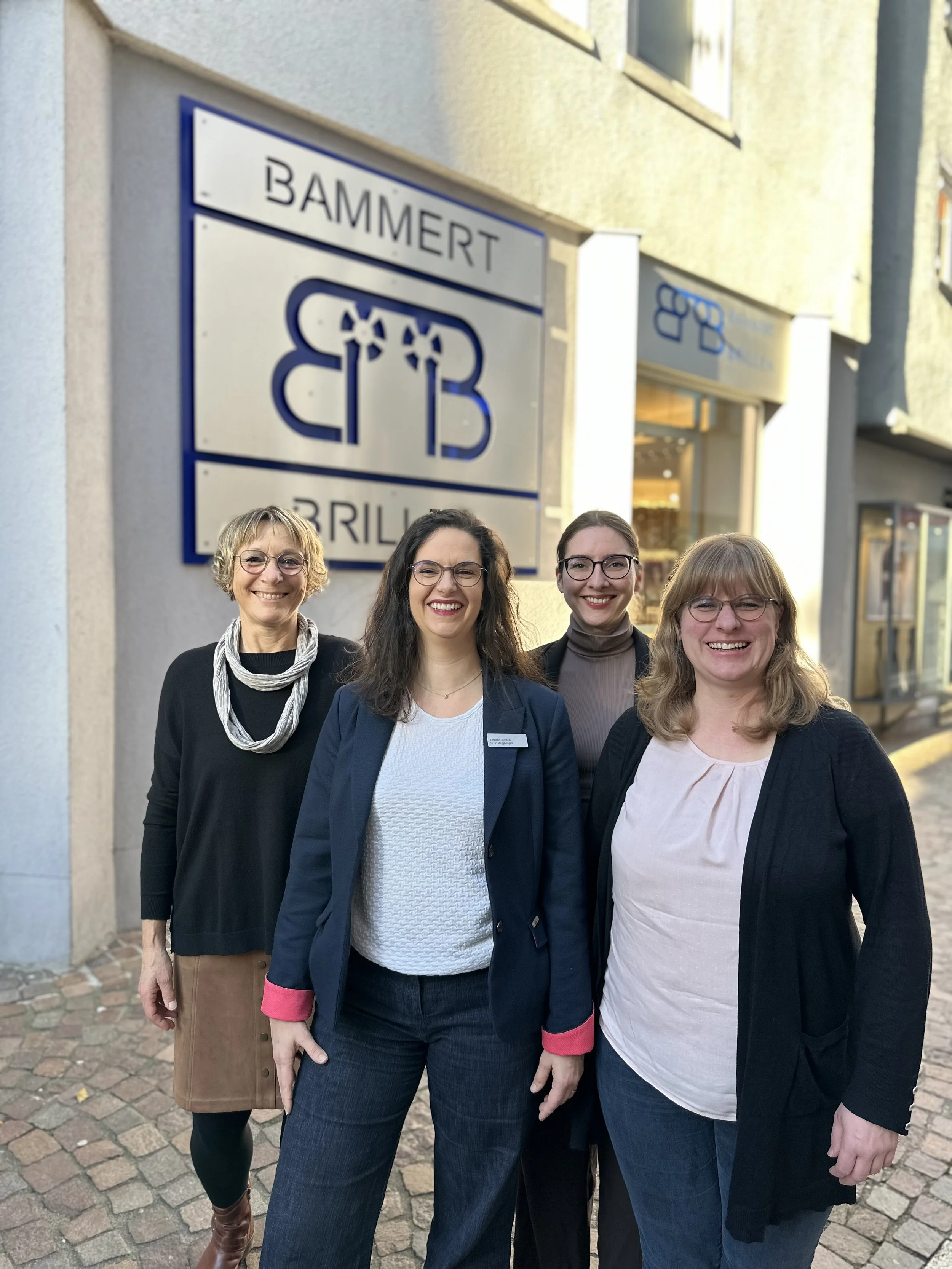 Vier Frauen stehen lächelnd vor einem Geschäft mit einem blauen und weißen Logo, das die Buchstaben „BB“ sowie den Namen „Bammert Brillen“ zeigt. Sie befinden sich auf einem gepflasterten Bürgersteig direkt vor dem Eingang des modernen Optikergeschäf