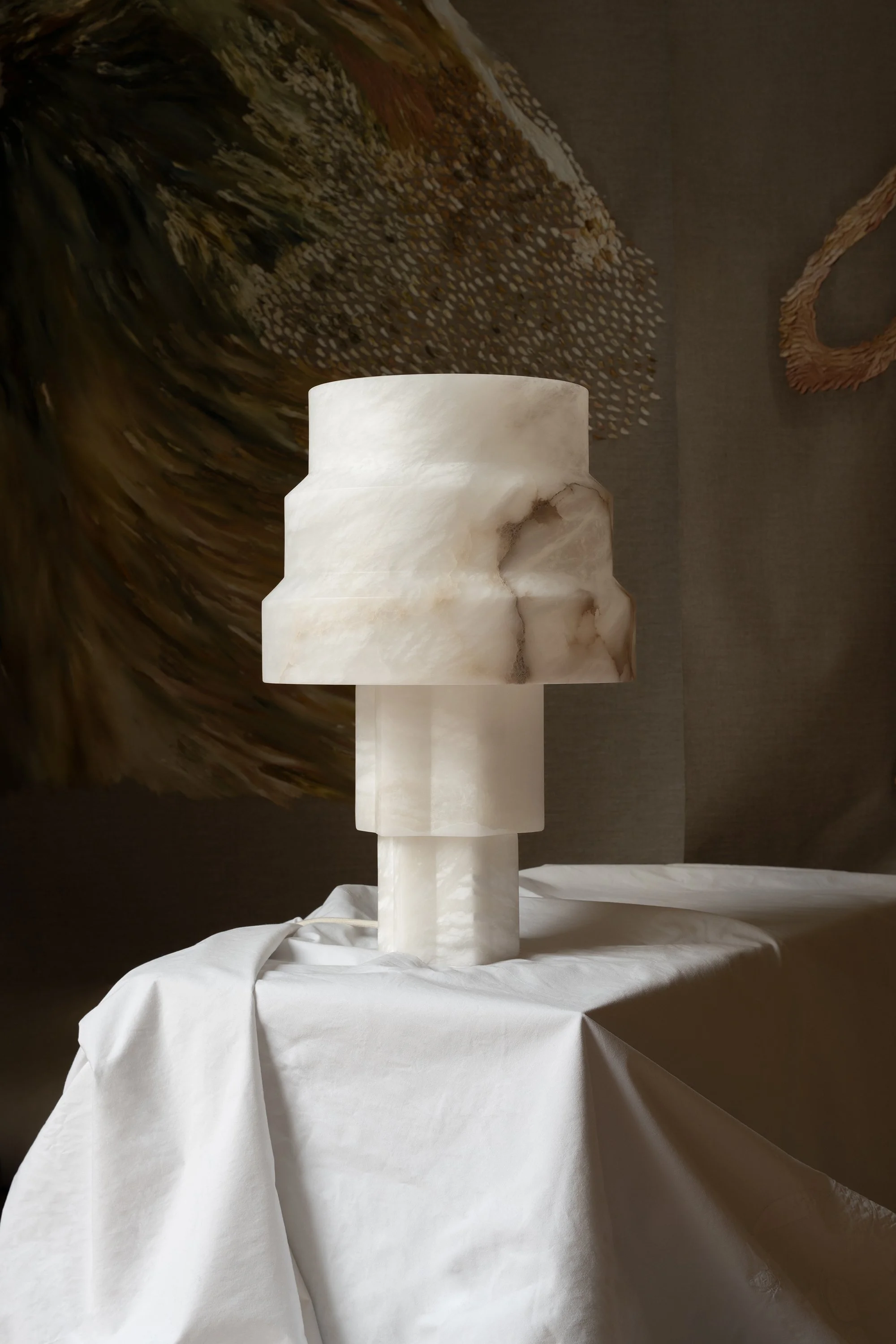 Une sculpture en pierre blanche représentant une forme géométrique avec plusieurs niveaux, placée sur une table drapée d'un tissu blanc, avec un fond décoratif abstrait.