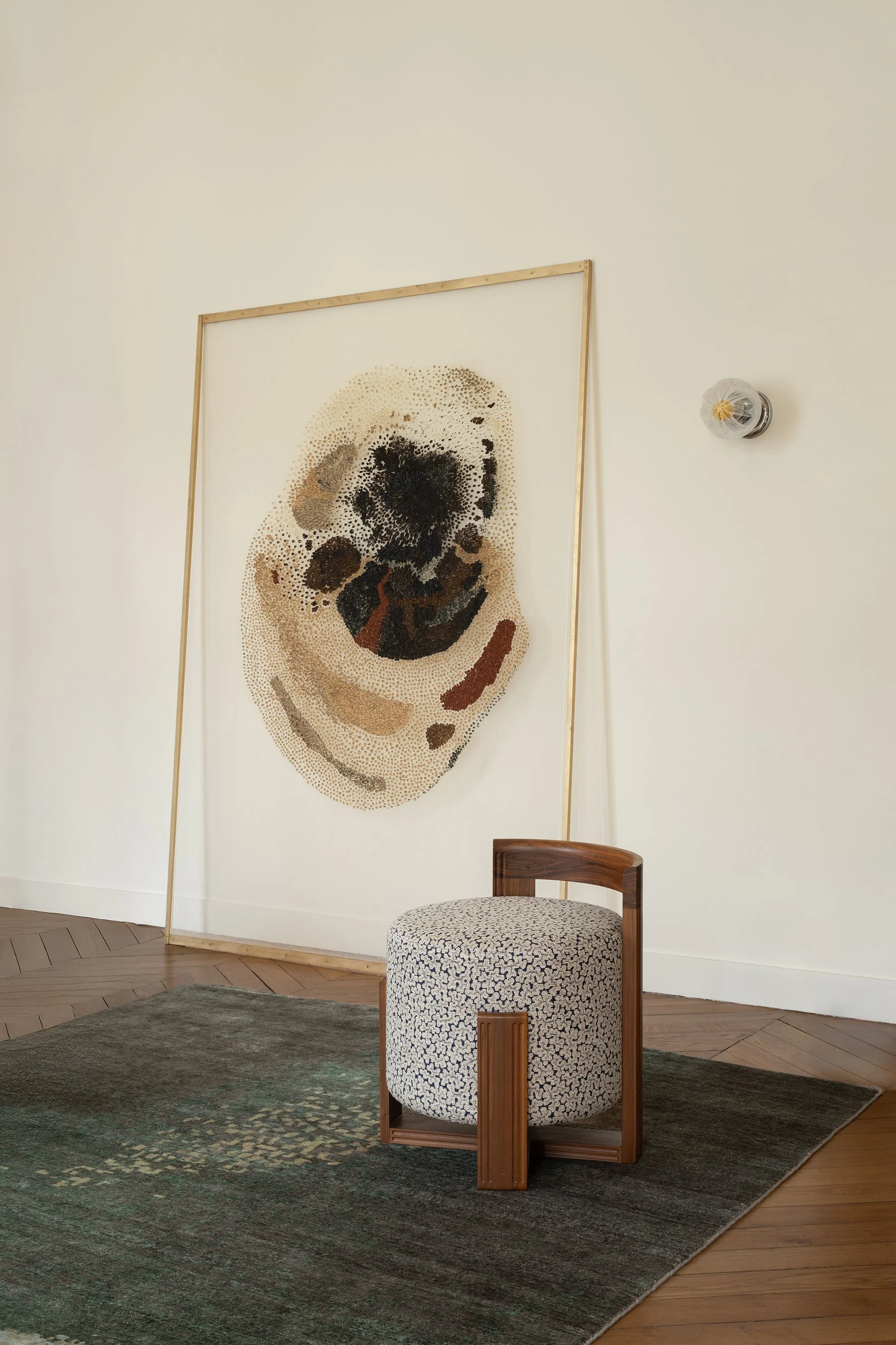 Une pièce d'art à portrait d'un chien réalisé avec de petits points, encadré en bois, placé derrière un tabouret avec assise en tissu à motif, sur un tapis vert foncé et un sol en bois, avec un mur beige et une applique murale en forme d'ampoule en arrière-plan.