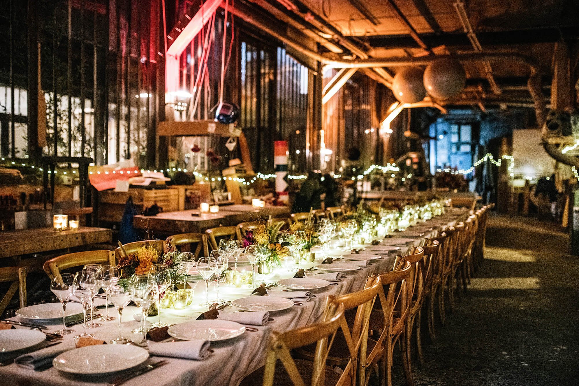 Une longue table est décorée pour un repas, avec des verres, assiettes, napperons, et centres de table floraux. La scène se déroule dans un espace intérieur ou semi-intérieur avec une ambiance chaleureuse, éclairée par des bougies et des guirlandes lumineuses, donnant une atmosphère festive ou de réception.