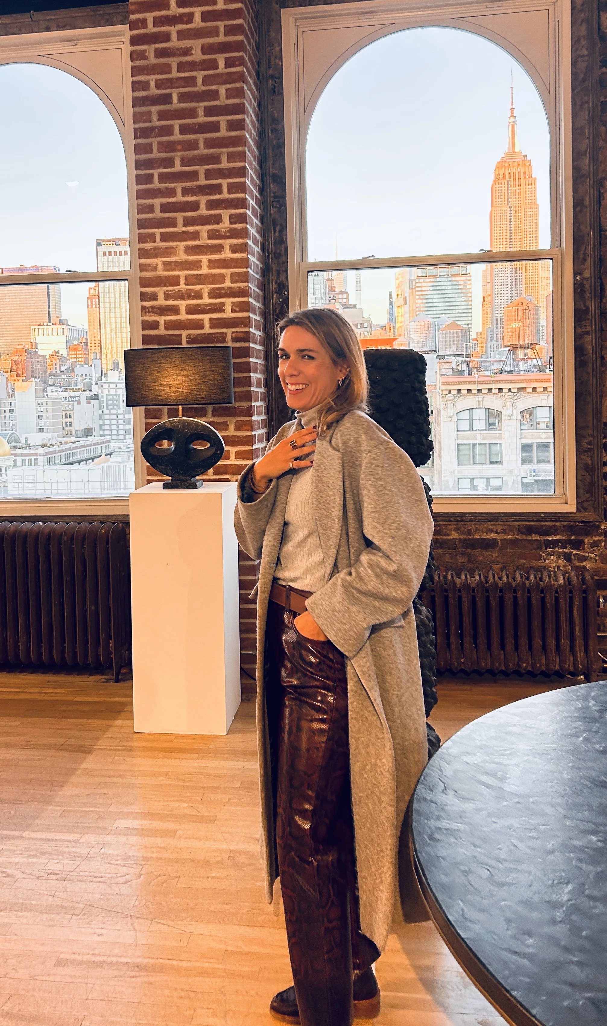 Femme souriante posant dans un appartement avec vue sur la skyline de New York, notamment l'Empire State Building, lors du coucher du soleil.