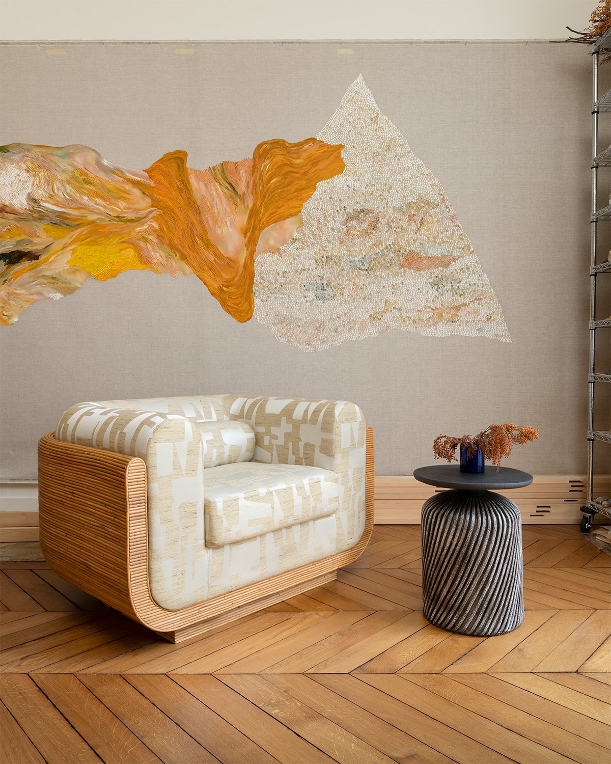 Un salon avec un canapé beige à motifs, une table ronde grise avec un vase bleu et des branches séchées, et un tableau mural abstrait aux tons chauds, dans une pièce au sol en bois clair.
