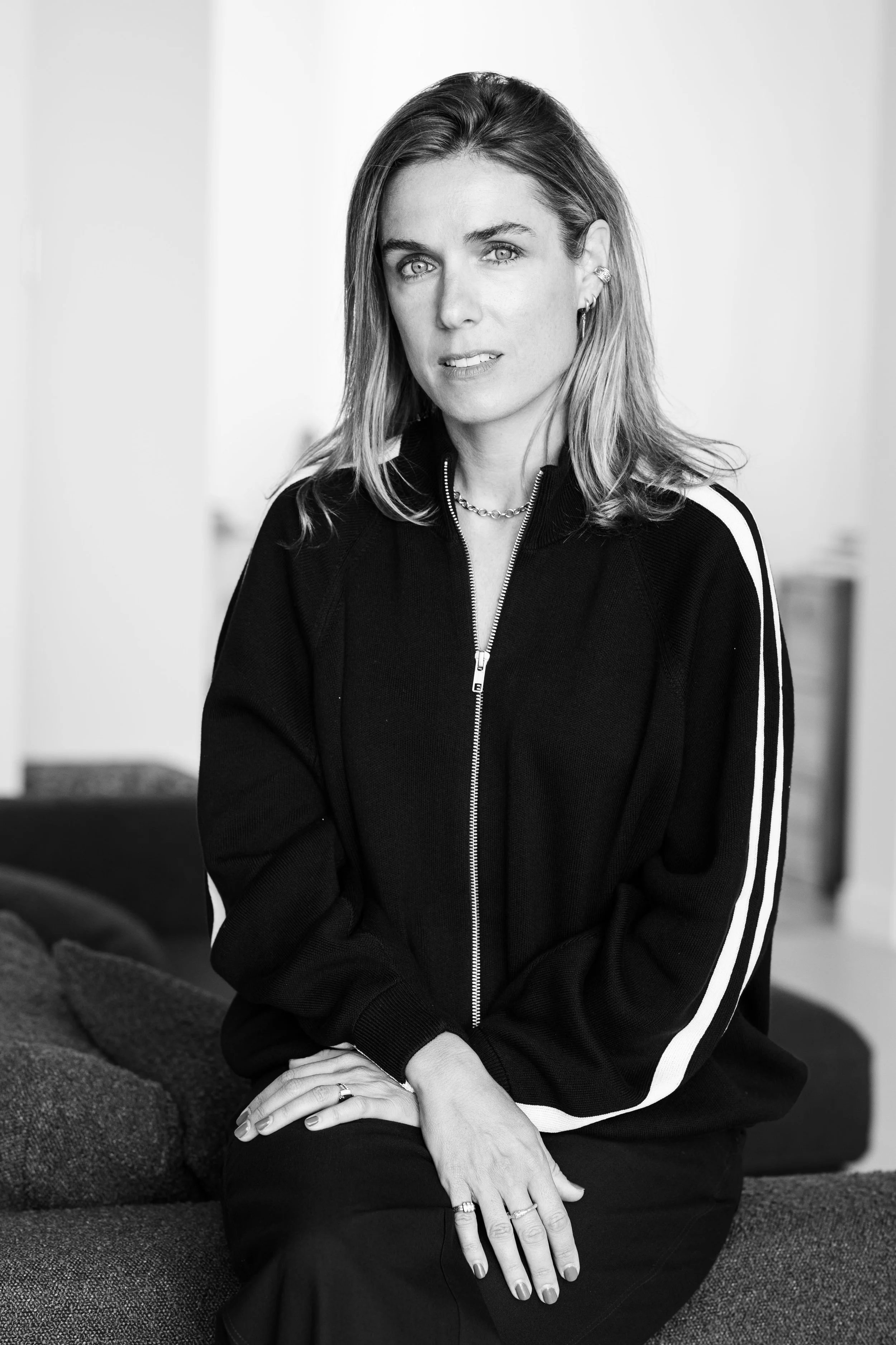 Portrait en noir et blanc d'une femme assise, portant un sweat à zip noir avec bandes blanches sur les manches, portant des bijoux et regardant directement l'objectif.
