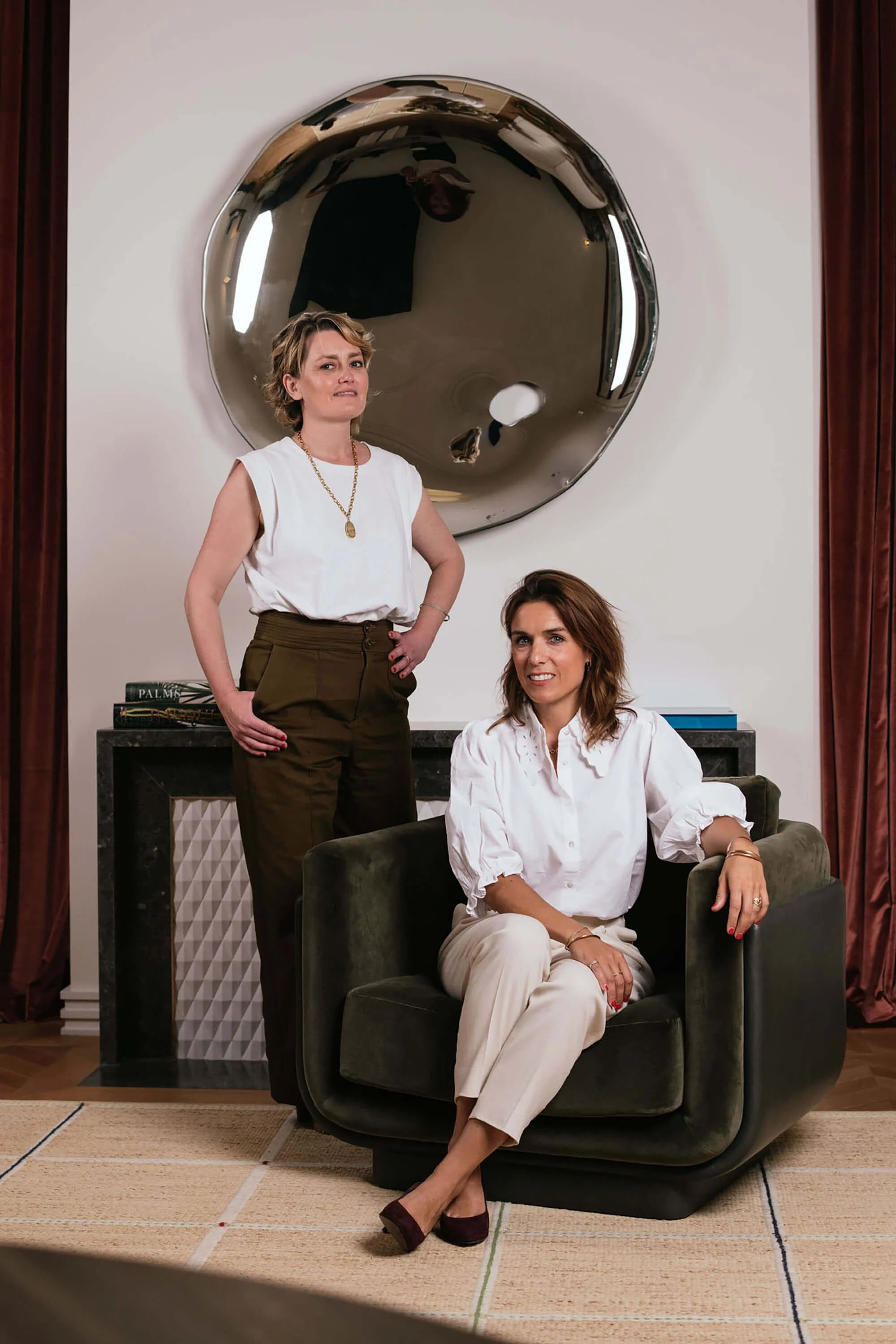 Deux femmes posent dans un salon, une debout avec une main sur la taille et l'autre assise sur un fauteuil, avec un grand miroir rond derrière elles.