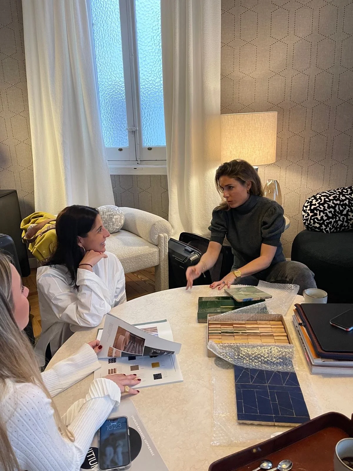 Deux femmes discutent lors d'une réunion à une table, entourées de matériaux de design d'intérieur, dans un salon avec une lampe, un canapé, et une fenêtre un peu givrée.