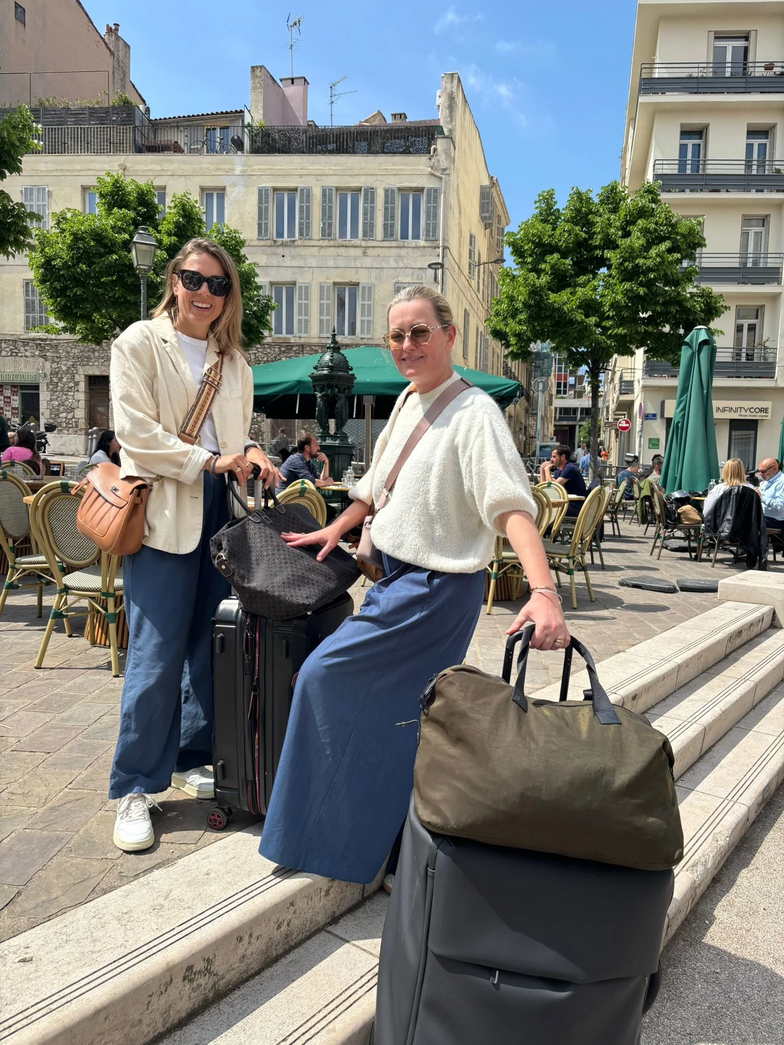 Deux femmes souriantes avec des bagages, assises sur un trottoir d'une place animée, avec des bâtiments résidentiels en arrière-plan, en plein jour avec un ciel bleu clair.