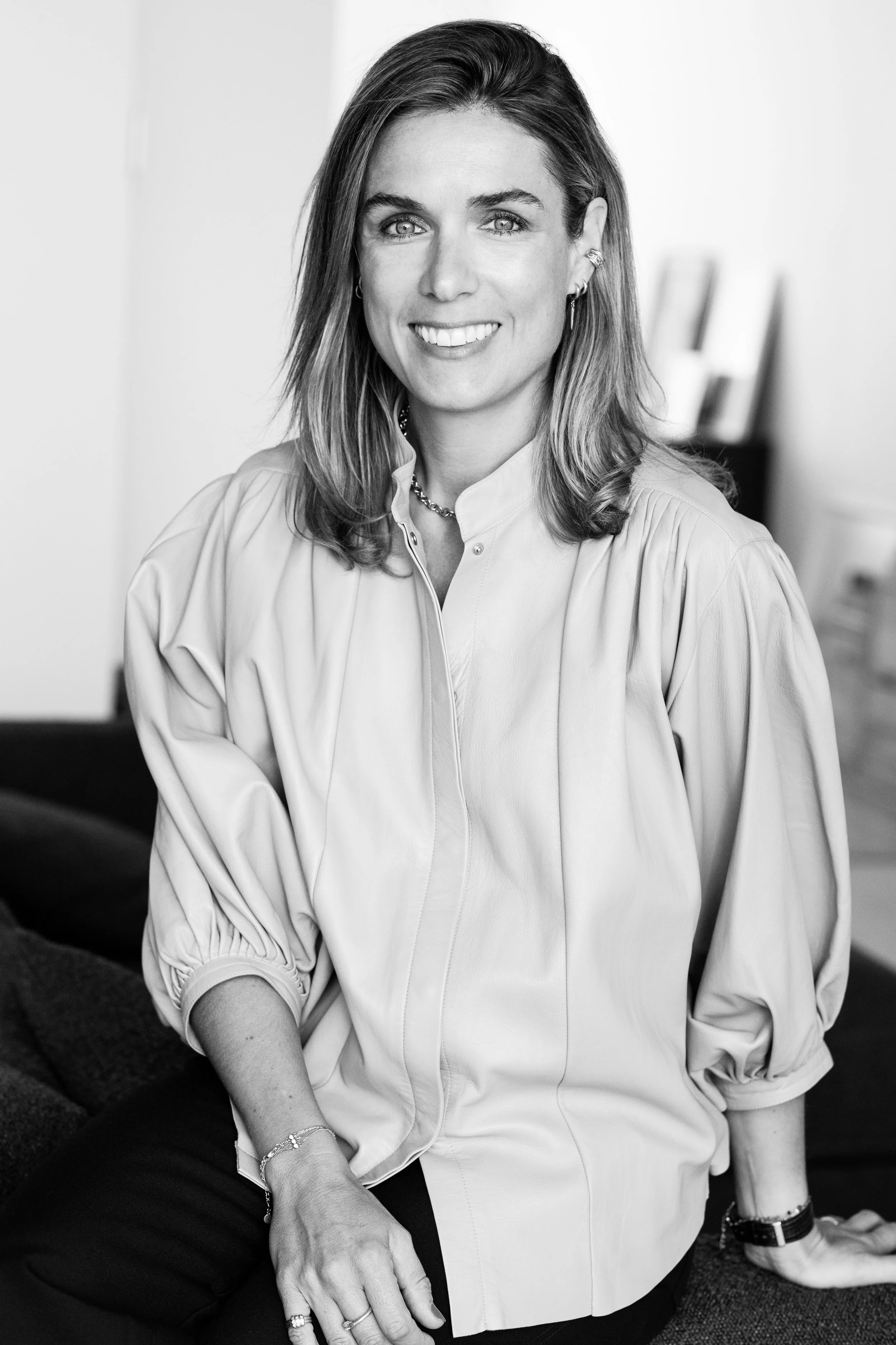 Portrait en noir et blanc d'une femme aux cheveux mi-longs, souriante, portant un chemisier clair, des boucles d'oreilles et un collier, assise dans un intérieur professionnel.