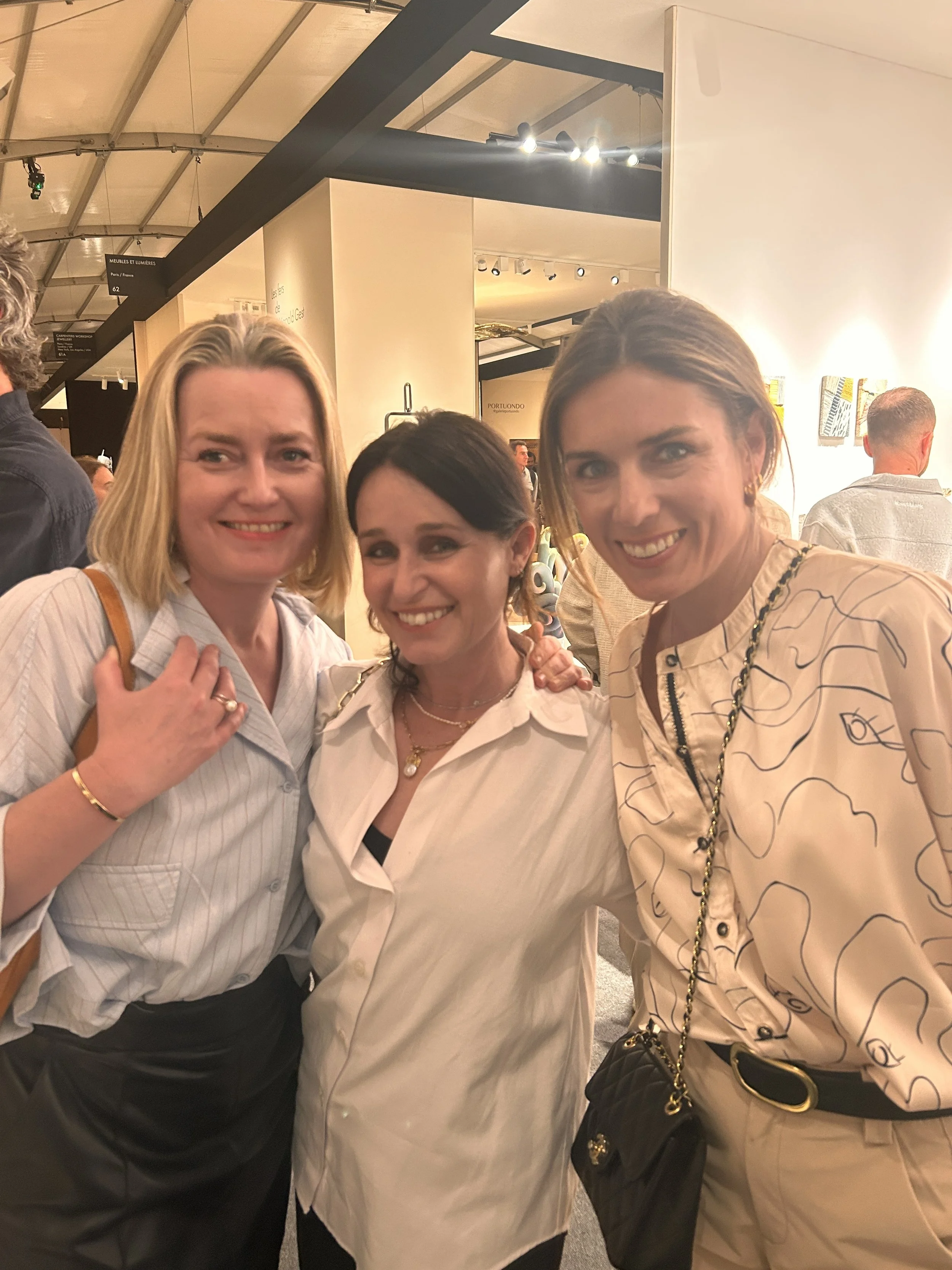 Trois femmes souriantes posent ensemble dans un lieu intérieur bien éclairé, avec des œuvres d'art visibles au mur à l'arrière.