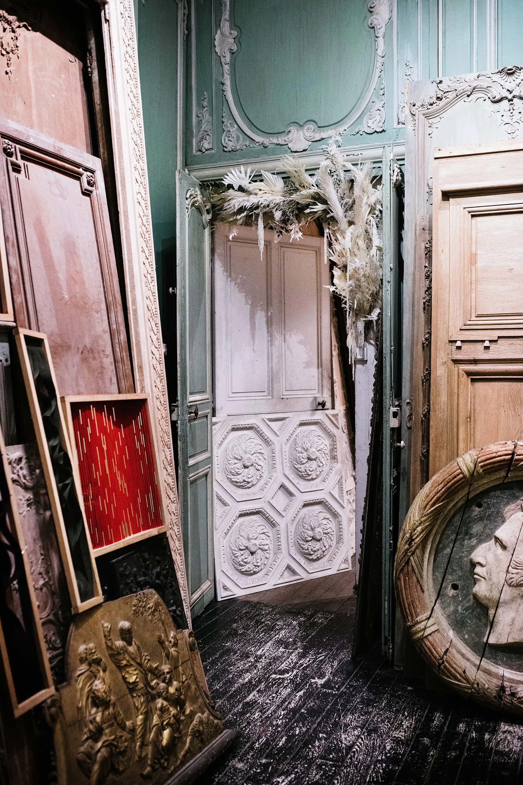 Une pièce avec des moulures ornées sur les murs et le plafond, plusieurs portes anciennes, des sculptures en bois et en plâtre, et des décorations artistiques, notamment une couronne en osier et une sculpture de visage romain.