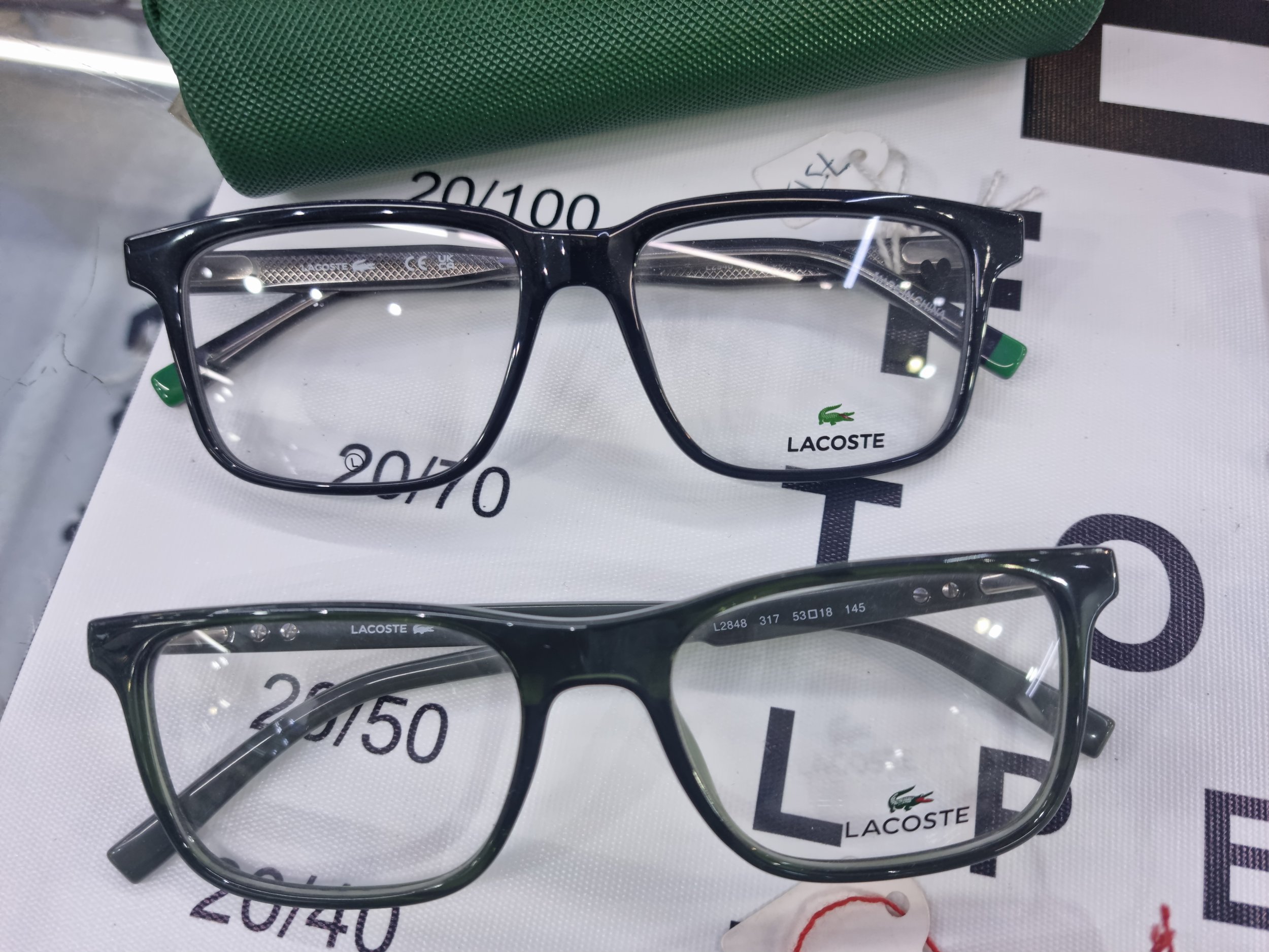 Two pairs of Lacoste eyeglasses on display, one above the other, on a white surface with Lacoste branding and measurement markings.