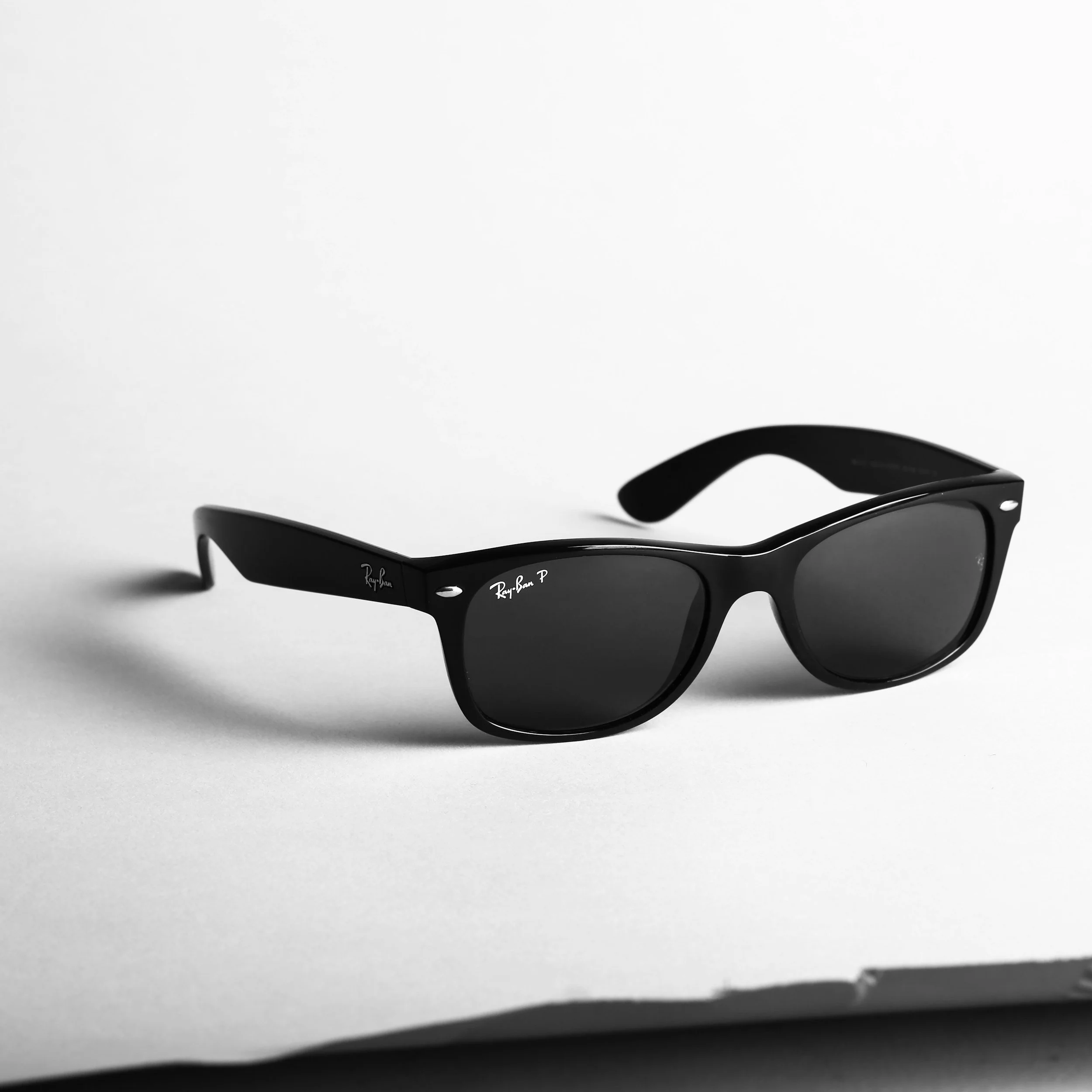 Black Ray-Ban sunglasses with dark lenses and white logo, resting on white paper background.