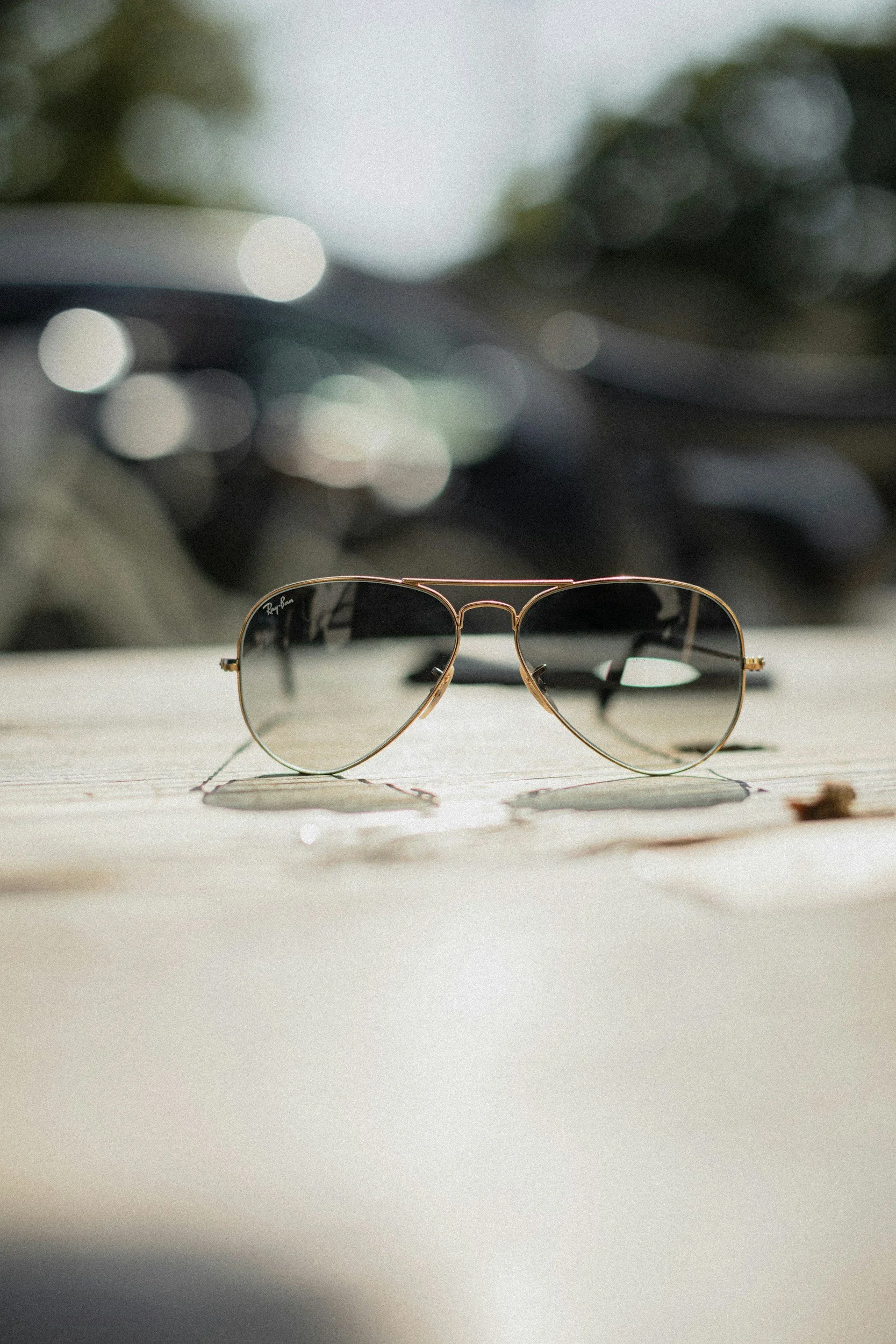 A pair of Ray-Ban sunglasses with dark lenses and a gold frame resting on a wooden surface outdoors.