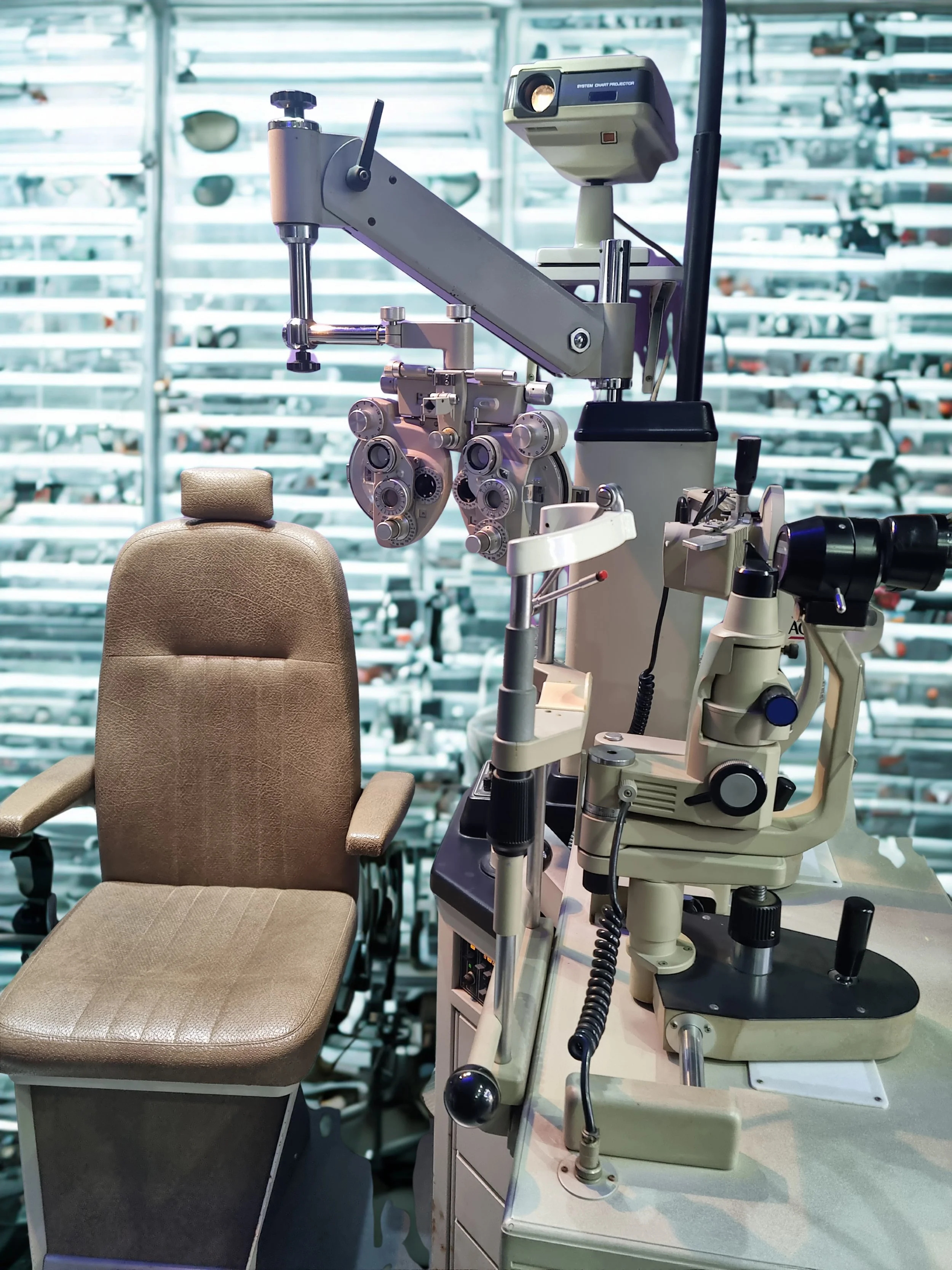 An ophthalmology exam room with a beige examination chair and a slit lamp microscope setup for eye examinations, against a background of stacked optical lenses.