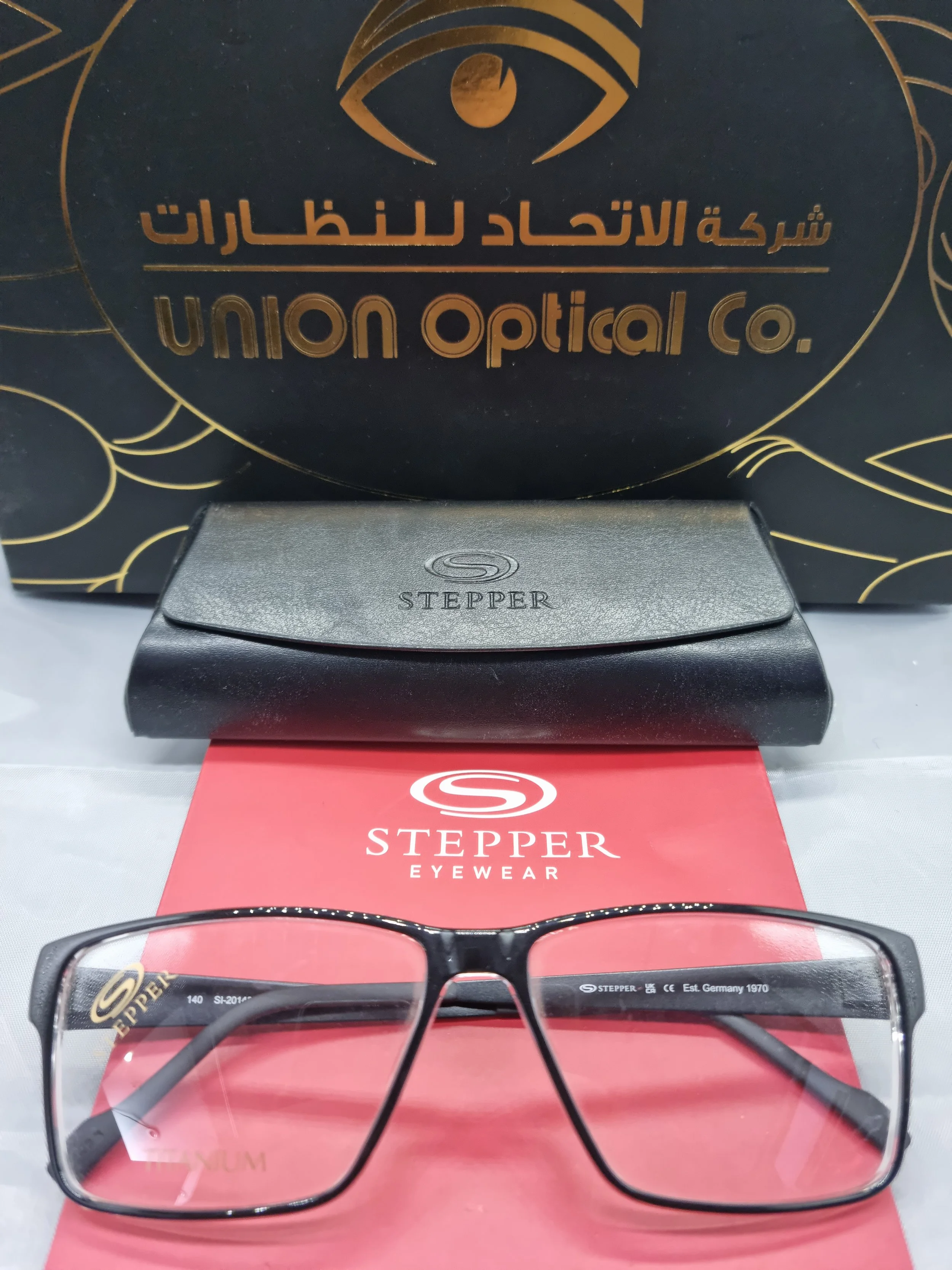 A pair of black and gray eyeglasses with the brand name 'Stepper' on the temples, placed on top of a red 'Stepper Eyewear' booklet; behind is a black plastic case with the 'Stepper' logo, and a black box with gold text and design featuring an eye log
