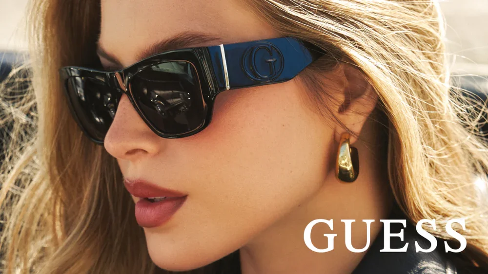 Close-up of a woman wearing black sunglasses with gold and blue details, gold earrings, and with light brown hair, outdoors in bright sunlight.