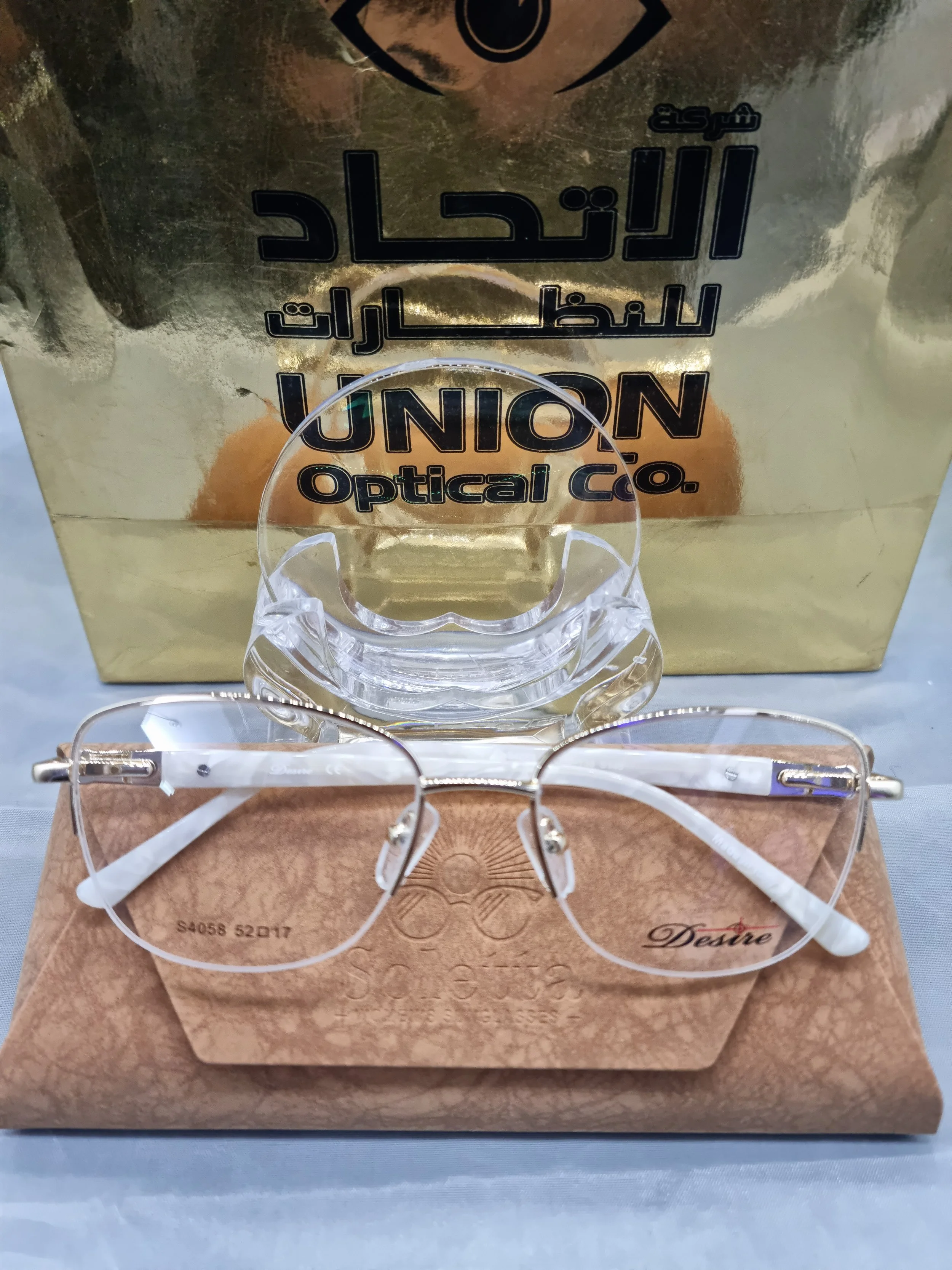 A pair of rimless eyeglasses with white temples resting on a brown textured case with the word 'Desire' written on it, placed in front of a gold-colored box labeled 'UNION Optical Co.'