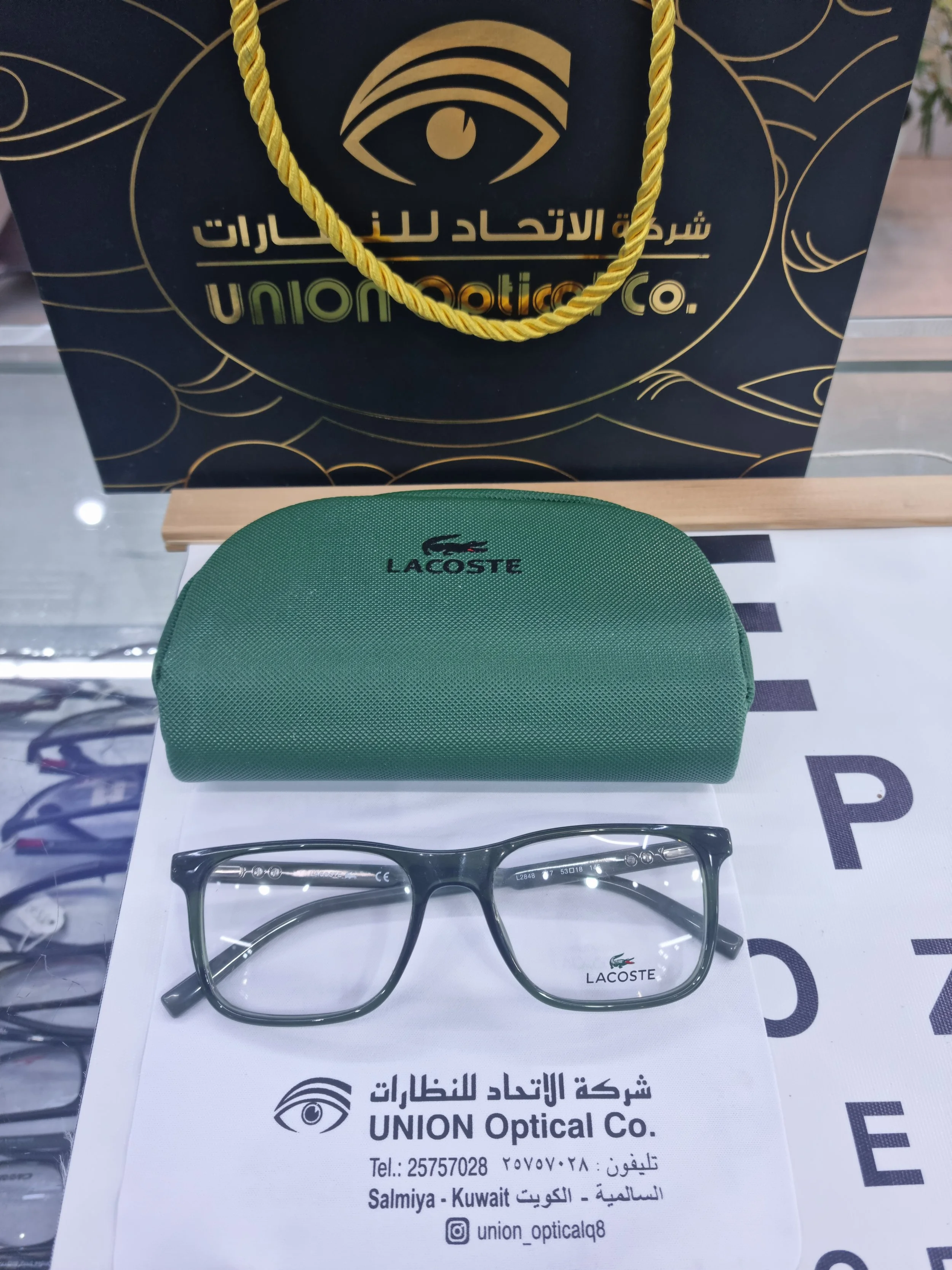A pair of black eyeglasses with clear lenses, a green Lacoste glasses case, and a white paper with store information. The background includes a Union Optical shopping bag with an eye logo and Arabic text.