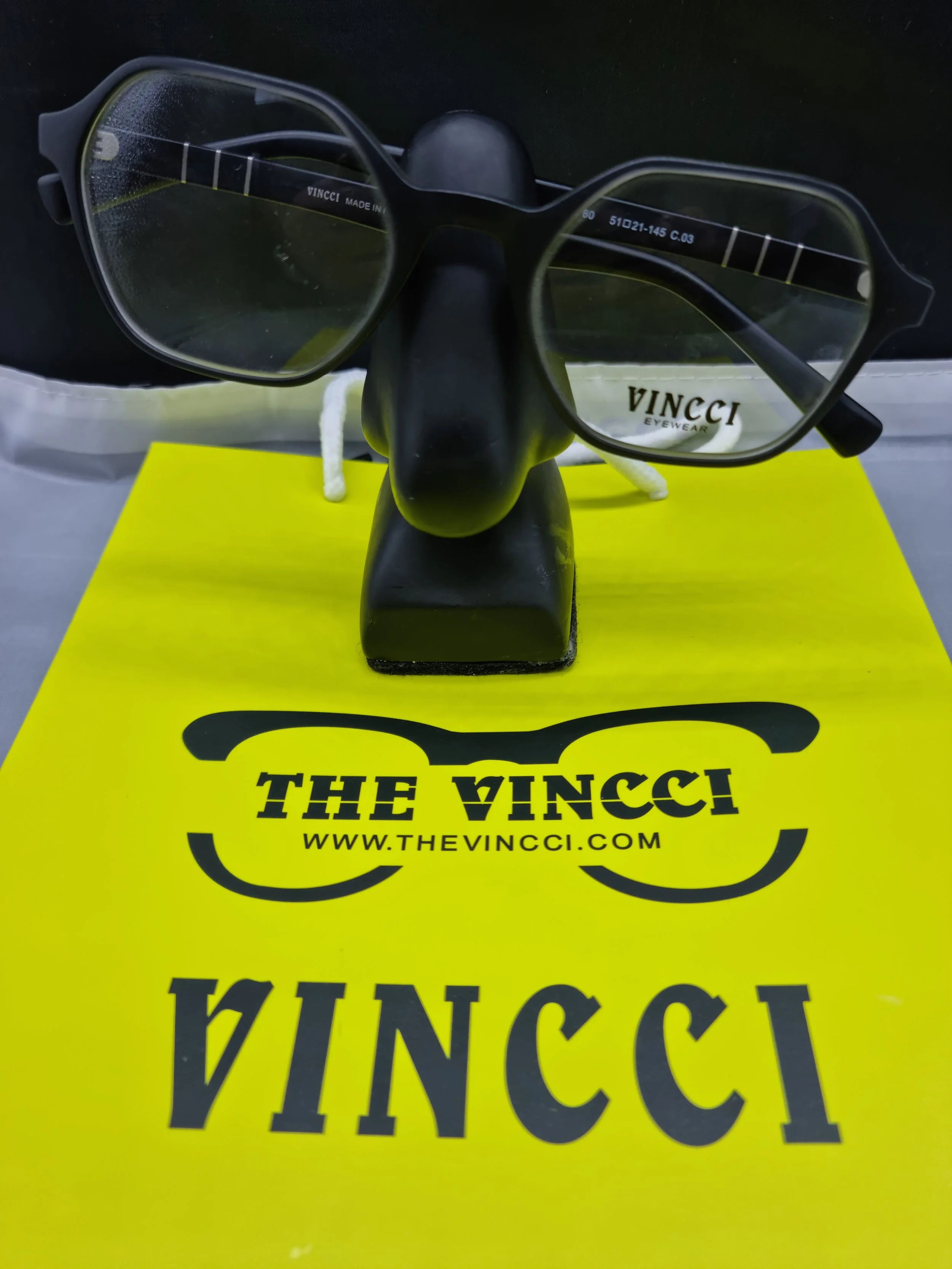 Pair of black eyeglasses displayed on a black stand, placed on a yellow surface with 'THE VINCCI' and a website URL printed on it.