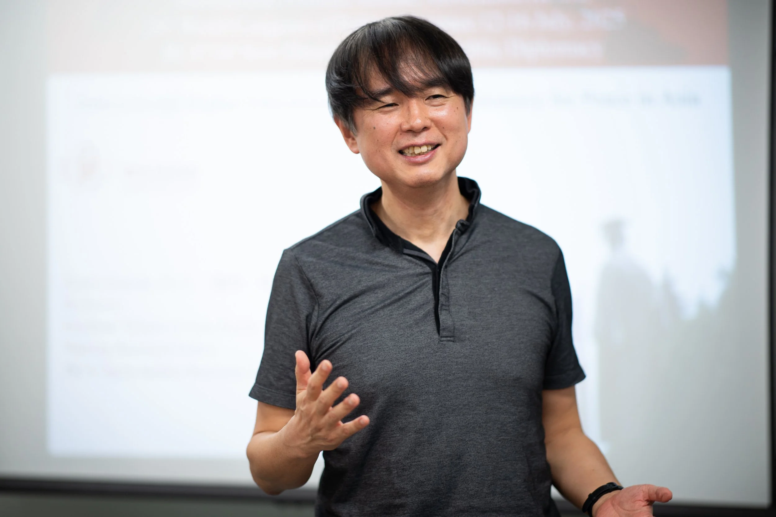 A man smiling and gesturing with one hand while standing in front of a presentation screen.