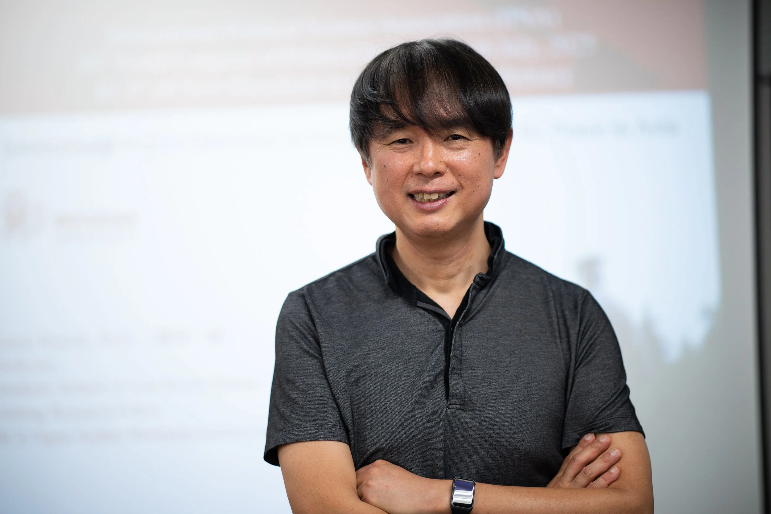 A man with black hair smiling and standing with arms crossed, wearing a black short-sleeved shirt and a fitness tracker on his left wrist, in front of a blurred background that appears to be a presentation slide.