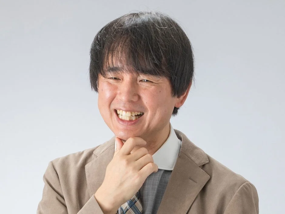 A man with black hair, smiling, wearing a beige blazer, gray shirt, and a striped tie, against a plain light background.