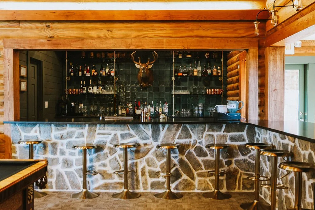 A rustic bar with a stone front, wooden beams, and a black countertop. Shelves behind hold liquor bottles and glassware, with a mounted deer head above.